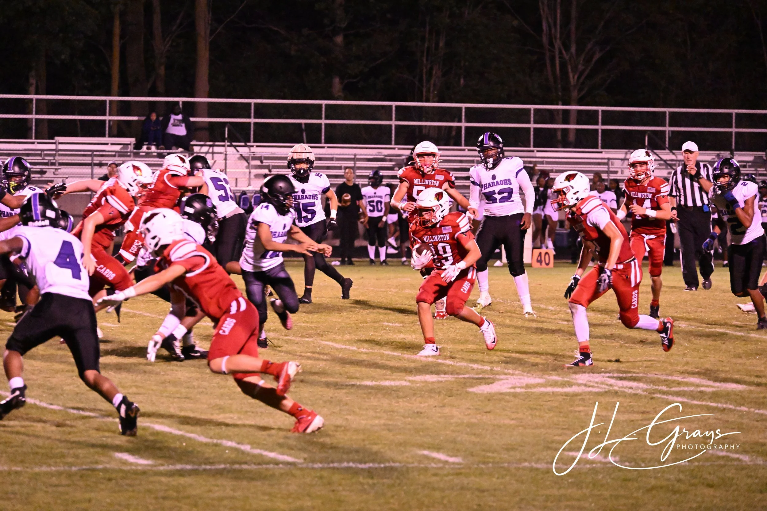 Young football players in red and white uniforms play a game on a grass field at night, with some players in white and others in red, with bleachers and trees in the background.