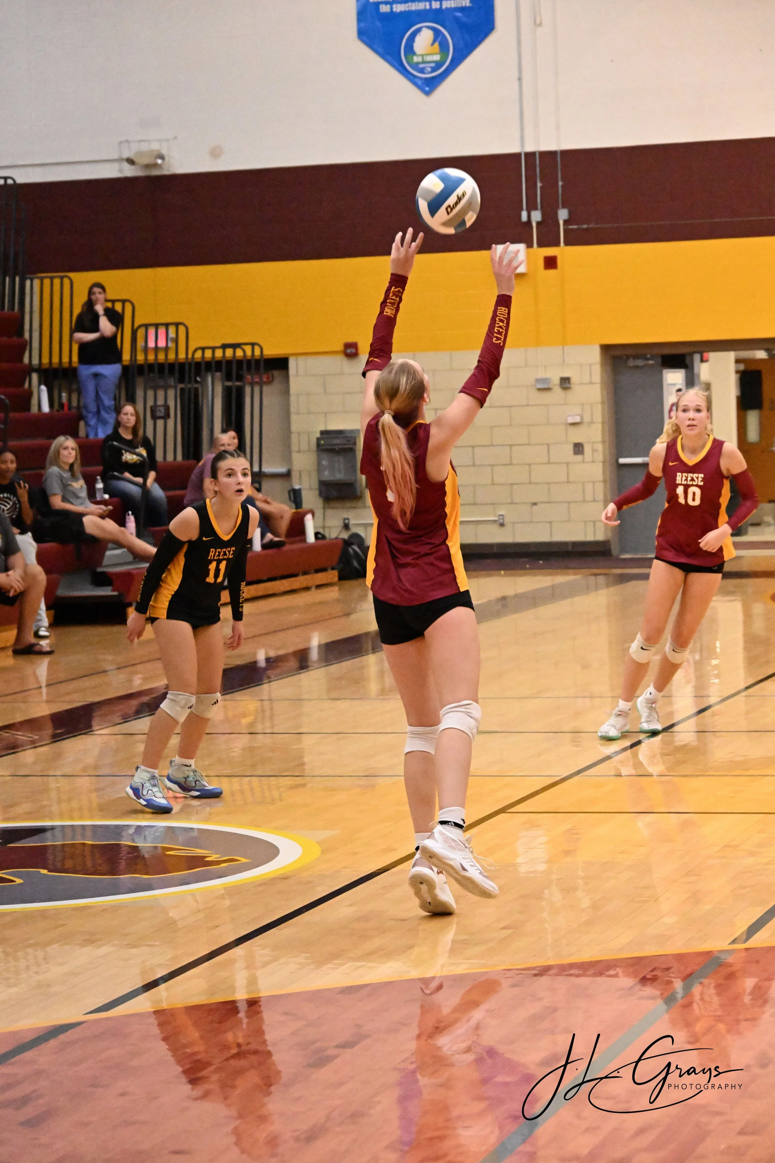 A volleyball game taking place in an indoor gymnasium. Three female players in black and red jerseys are visible, with one jumping to hit the ball. Spectators are sitting in the bleachers in the background.