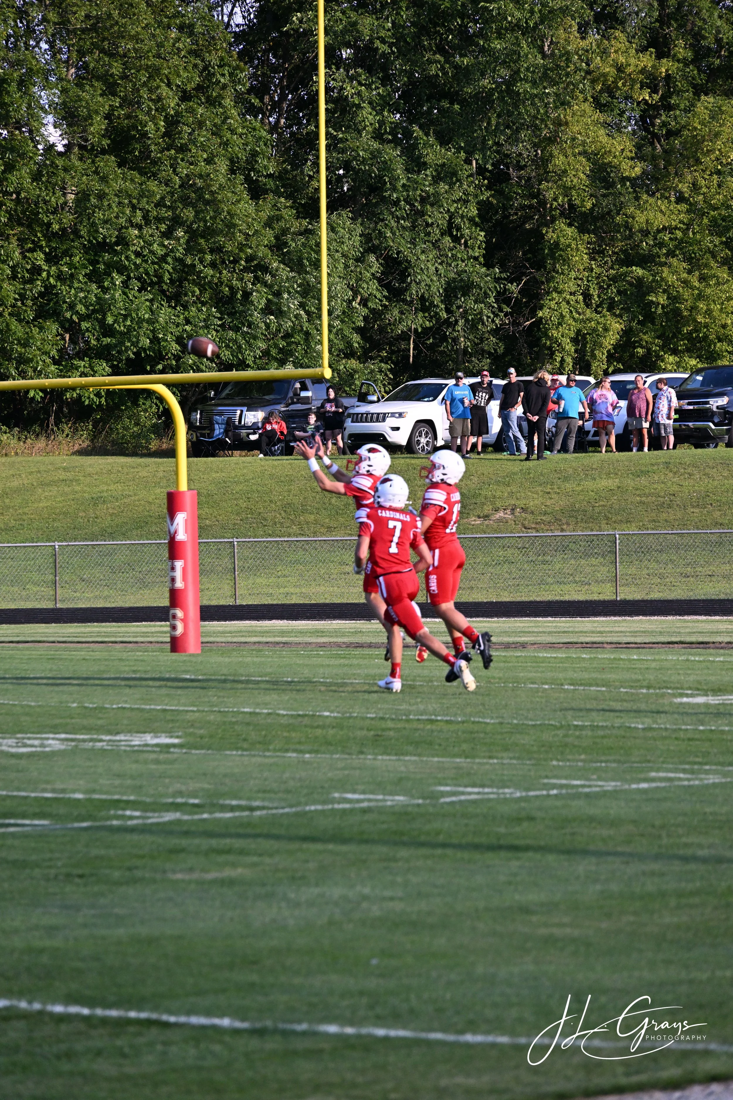American football players in red uniforms on a football field, with a football in mid-air near the goalpost, and spectators watching from the sidelines with cars parked in the background, surrounded by trees.