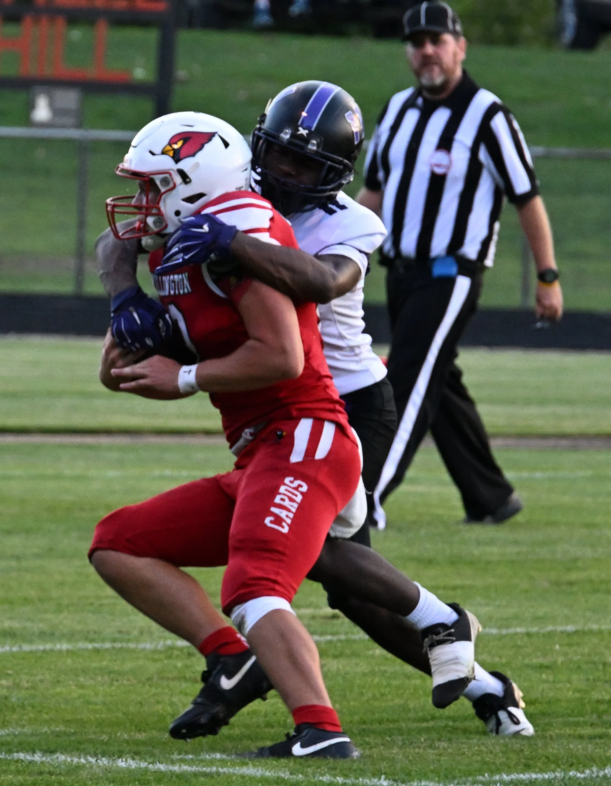 Football players in a game, one in a red uniform and helmet with a bird logo, running with the ball, while another in a white uniform and black helmet attempts to tackle him. An official in black and white striped shirt is in the background.
