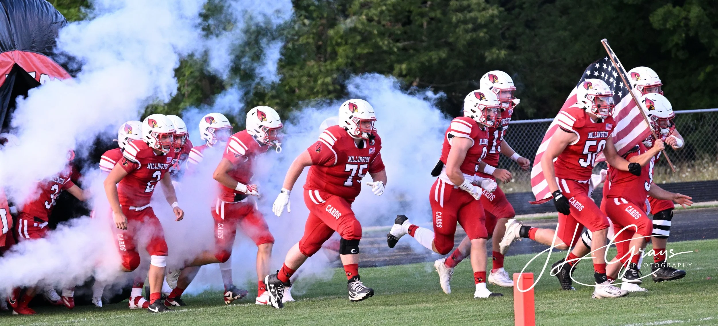 American football team in red uniforms running onto the field through smoke, with a player carrying an American flag.