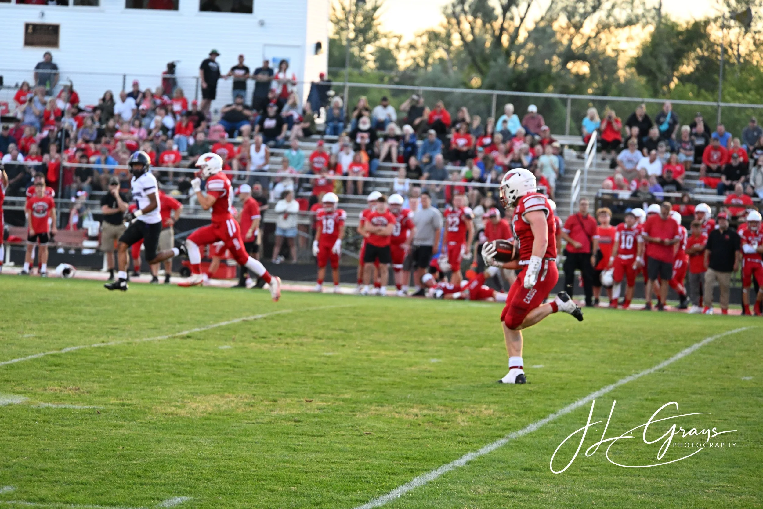 A football game at a stadium with players in red and white uniforms, some standing near the sideline, and spectators watching from the bleachers during the evening.