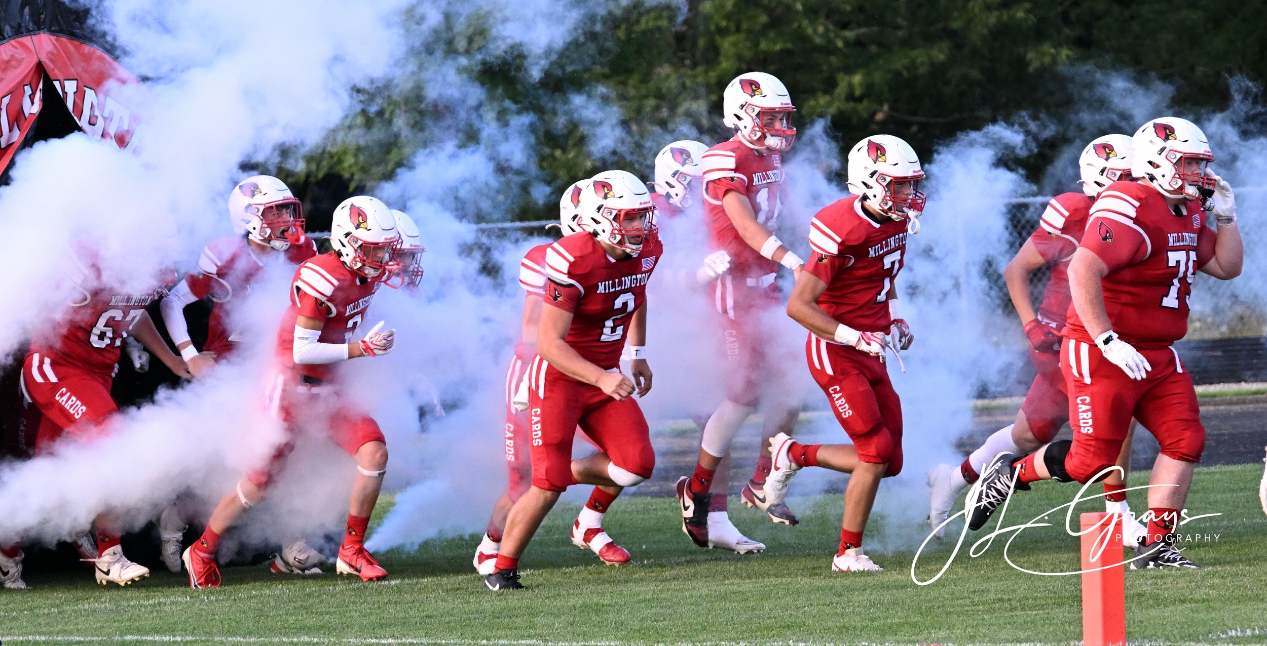 A football team in red uniforms, with some players running onto the field through smoke or mist, likely during a game or practice. The players are wearing helmets and pads, and the team name 'Millington' is visible on their jerseys. The background is