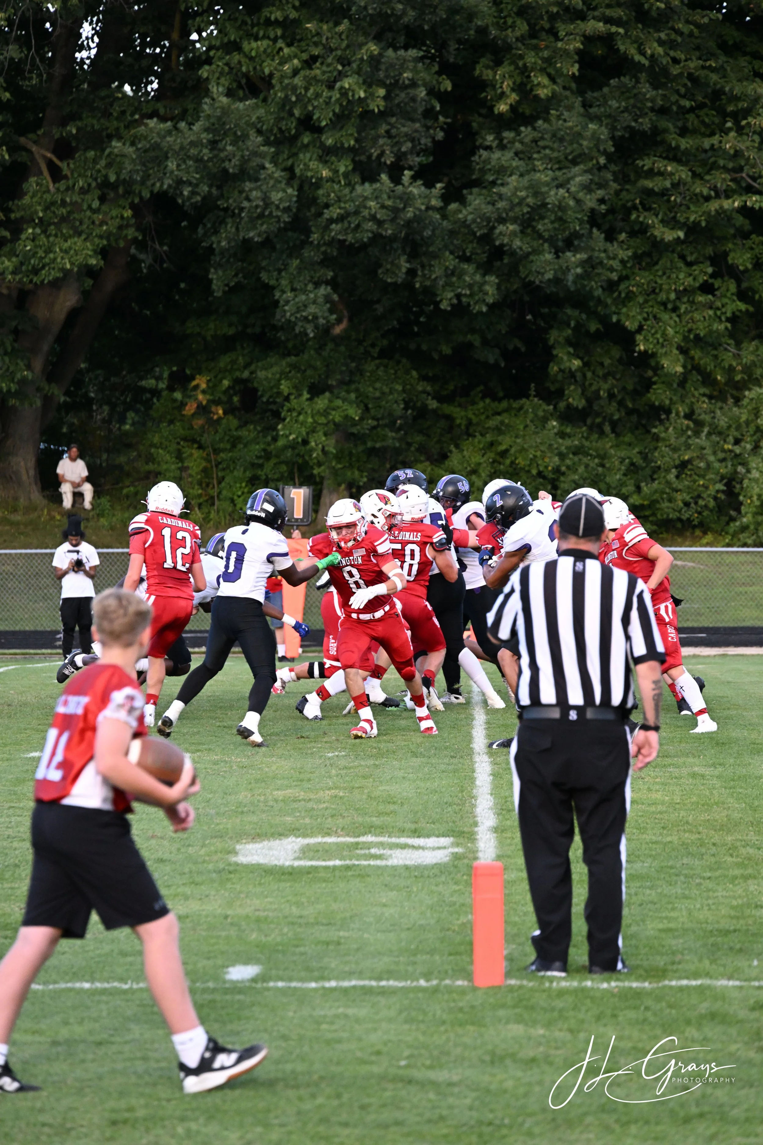 Young football players in red and white uniforms play a game on the field, with referees watching. Trees are visible in the background.