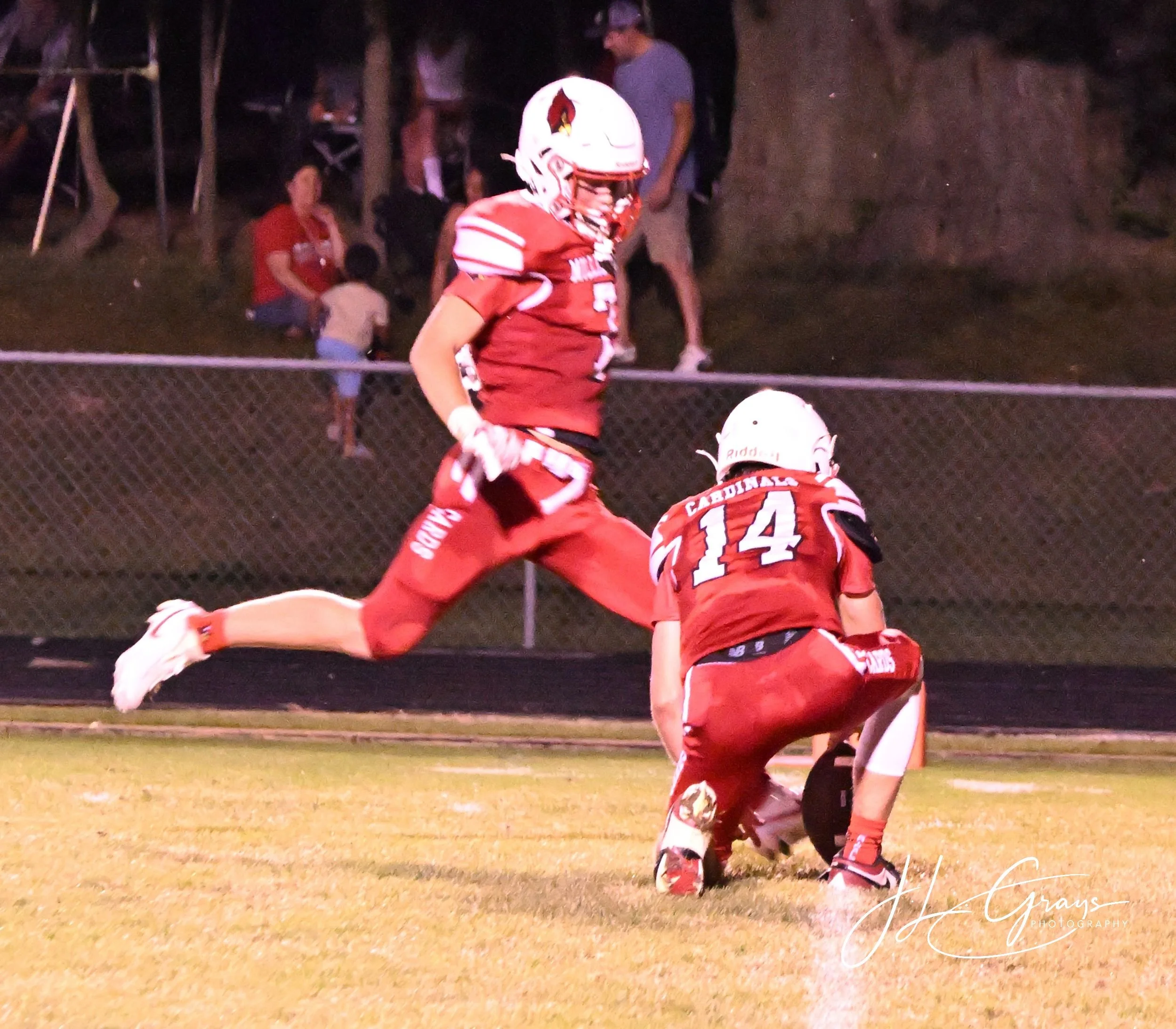 A football player in a red uniform and white helmet, numbered 14, kneeling on the field during a night game, with spectators watching from the sideline.
