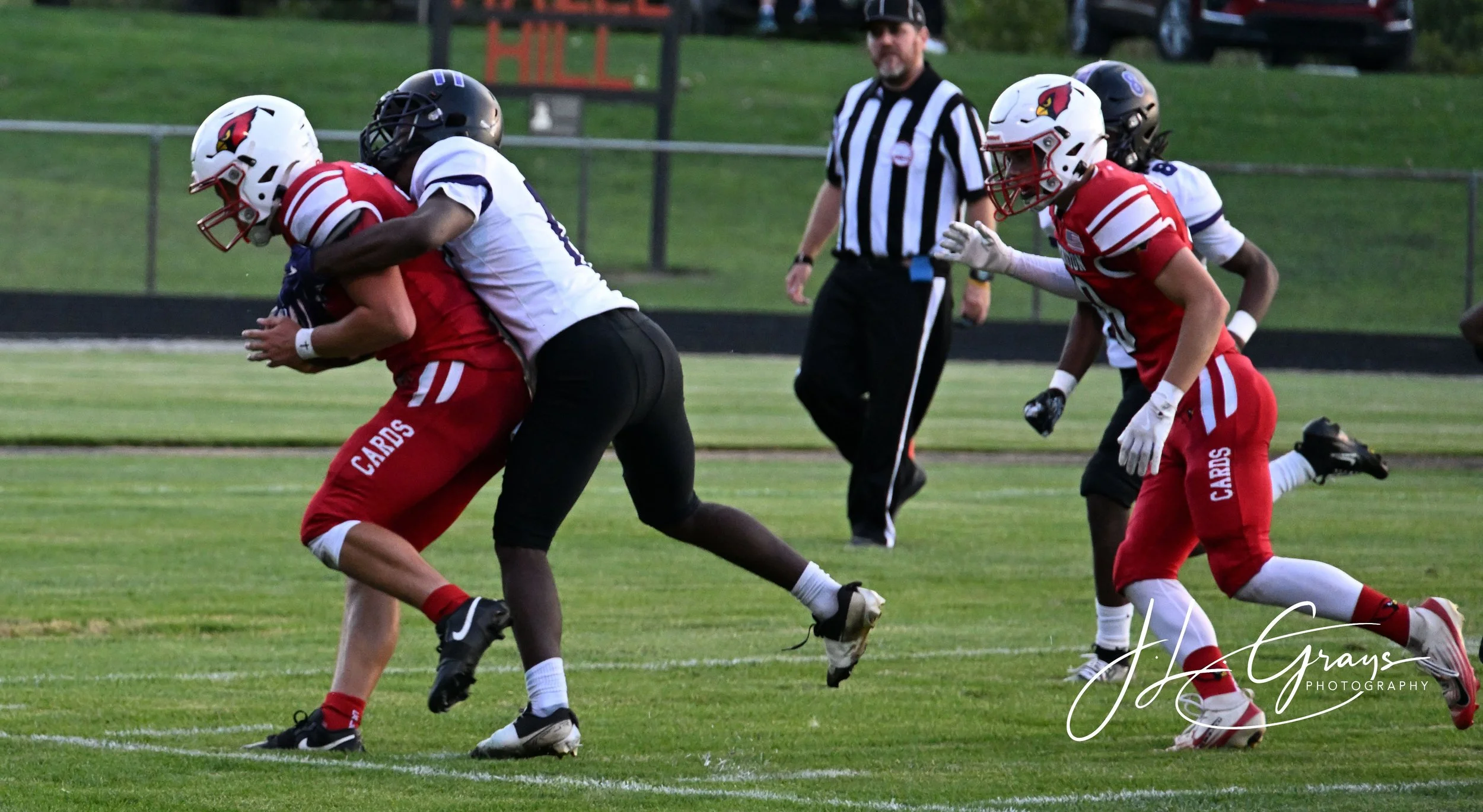 A football game with players in red and white uniforms tackling a player in a white uniform carrying the ball. An referee in black and white stripes observes the play on the field.