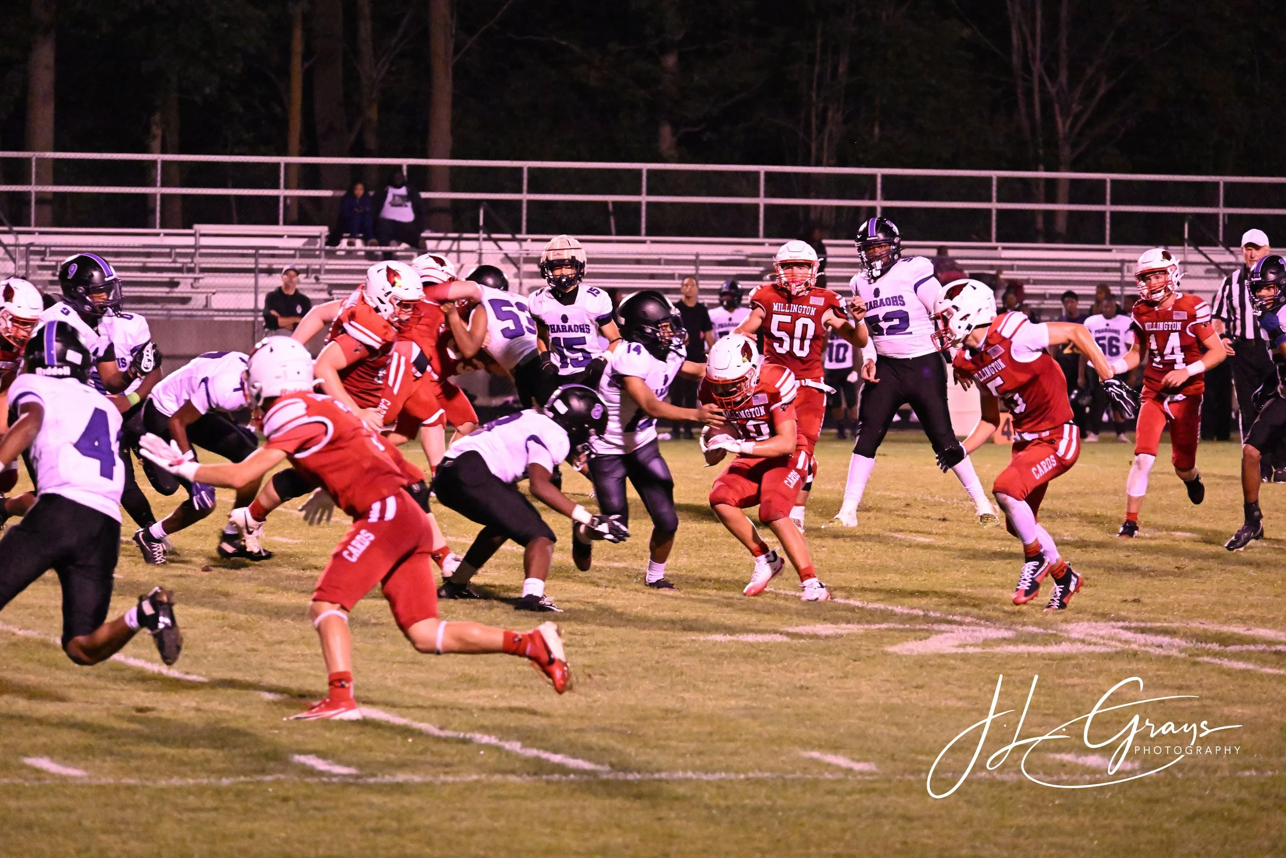 Photo of a football game at night, players in red and white uniforms tackle players in purple and white uniforms on a grassy field, with empty bleachers and trees in the background.