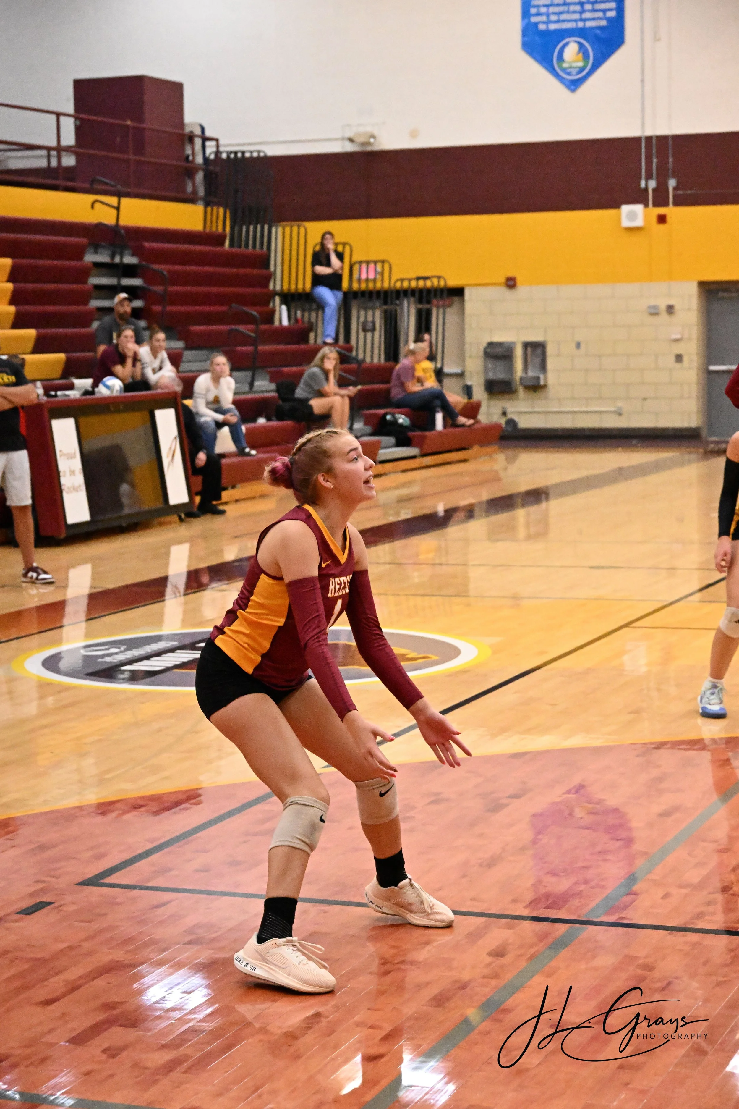 A female volleyball player with red hair in a pink bun, wearing a maroon and orange jersey, white knee pads, and white shoes, is in a ready stance on a volleyball court. In the background, a few spectators are seated in bleachers and watching the gam