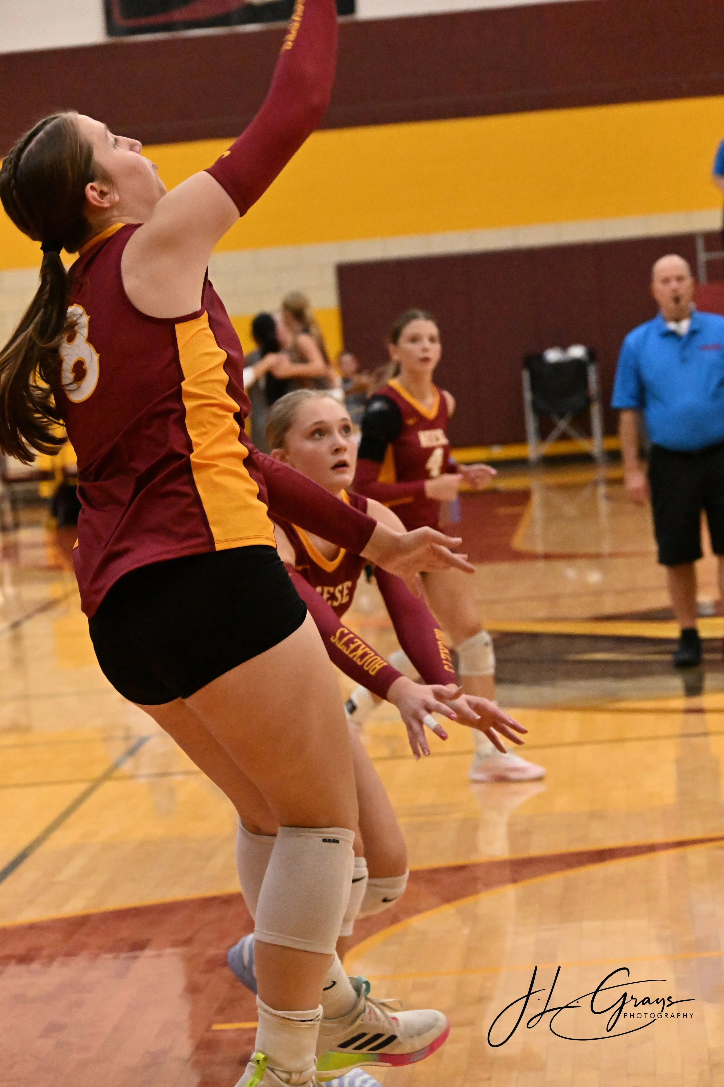 A volleyball player in a maroon and yellow uniform is preparing to serve during a game in a gymnasium with a yellow and maroon wall. Other players and a referee are visible in the background.