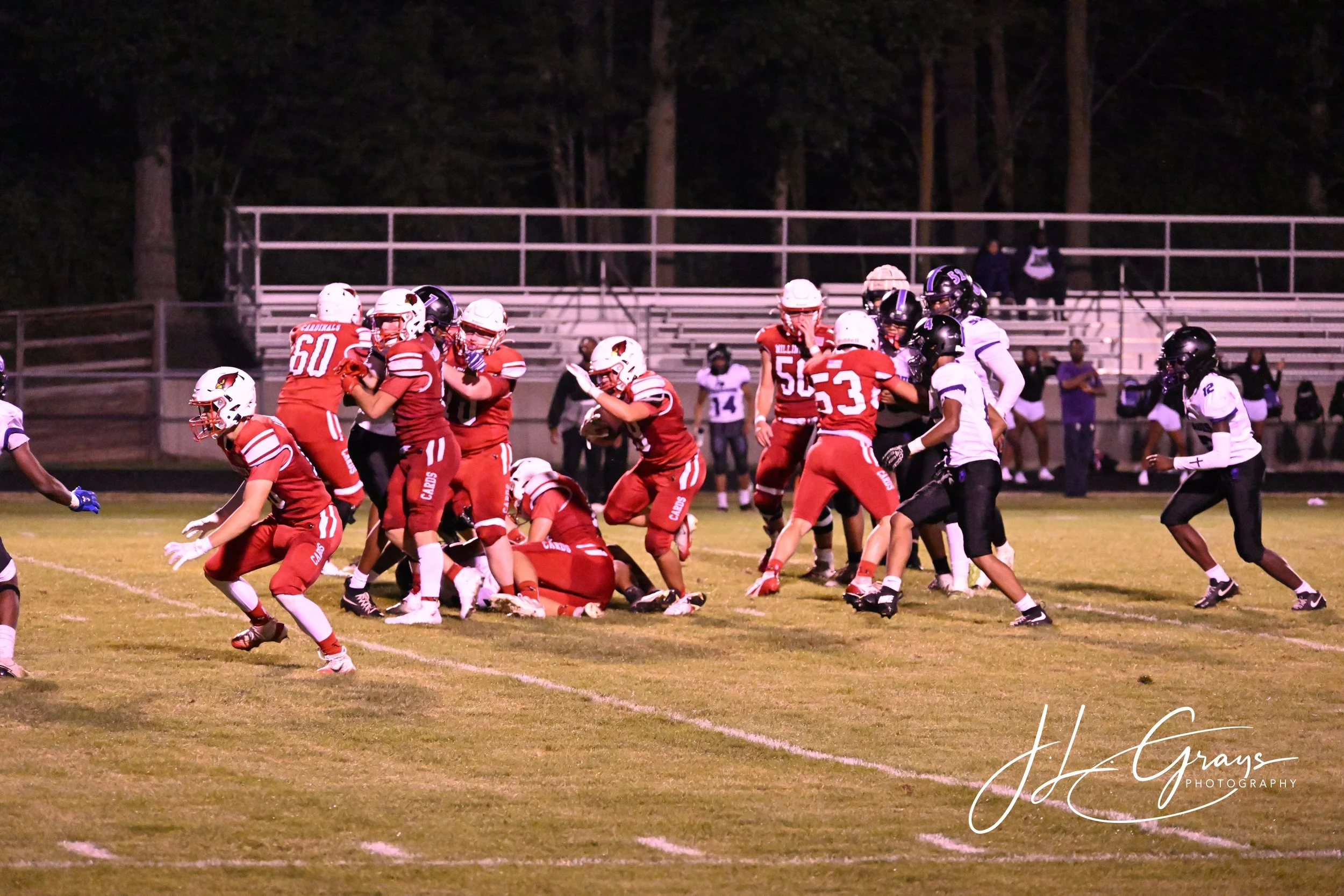 A nighttime football game with players on the field, some in red uniforms and others in black and white uniforms, engaged in a play. The background shows empty bleachers and trees.