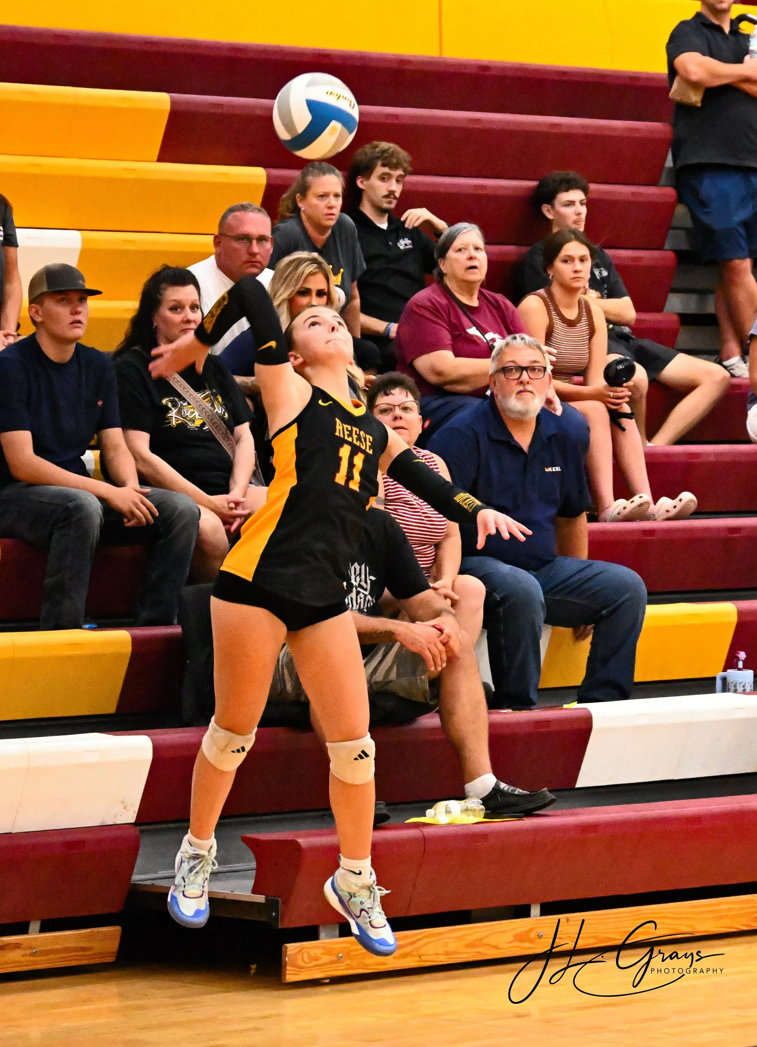 A female volleyball player in black and yellow uniform with the number 11, preparing to serve, on a gymnasium court with spectators seated on red and yellow bleachers.