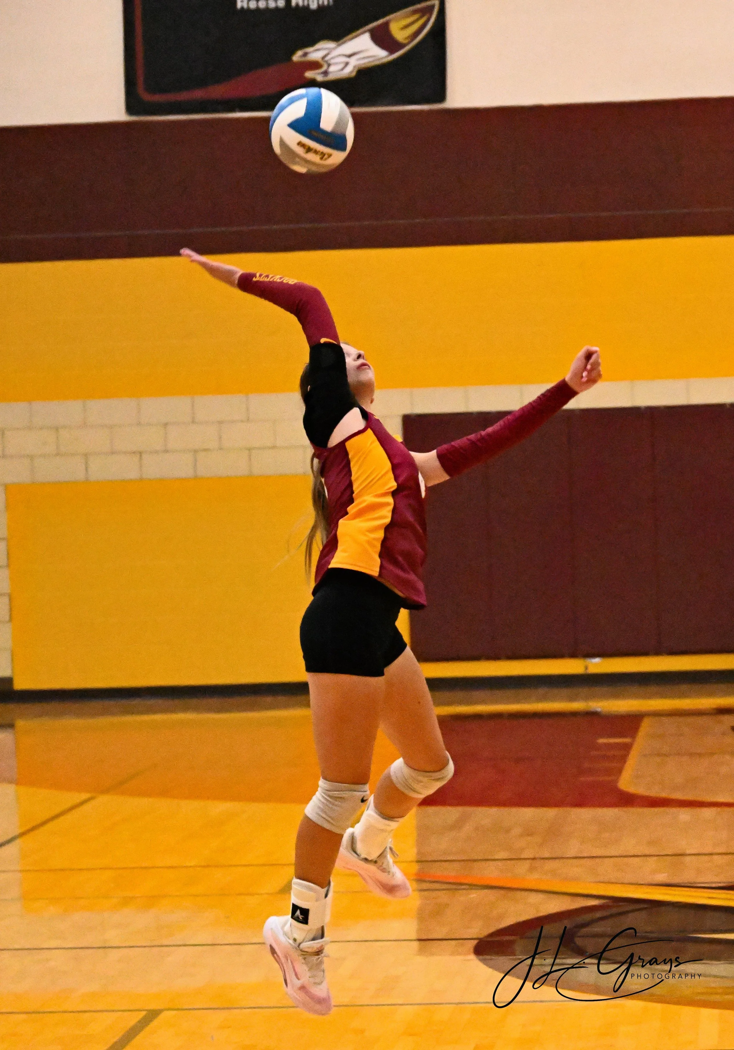 A female volleyball player wearing a maroon and yellow uniform jumps to hit a volleyball in an indoor gym.