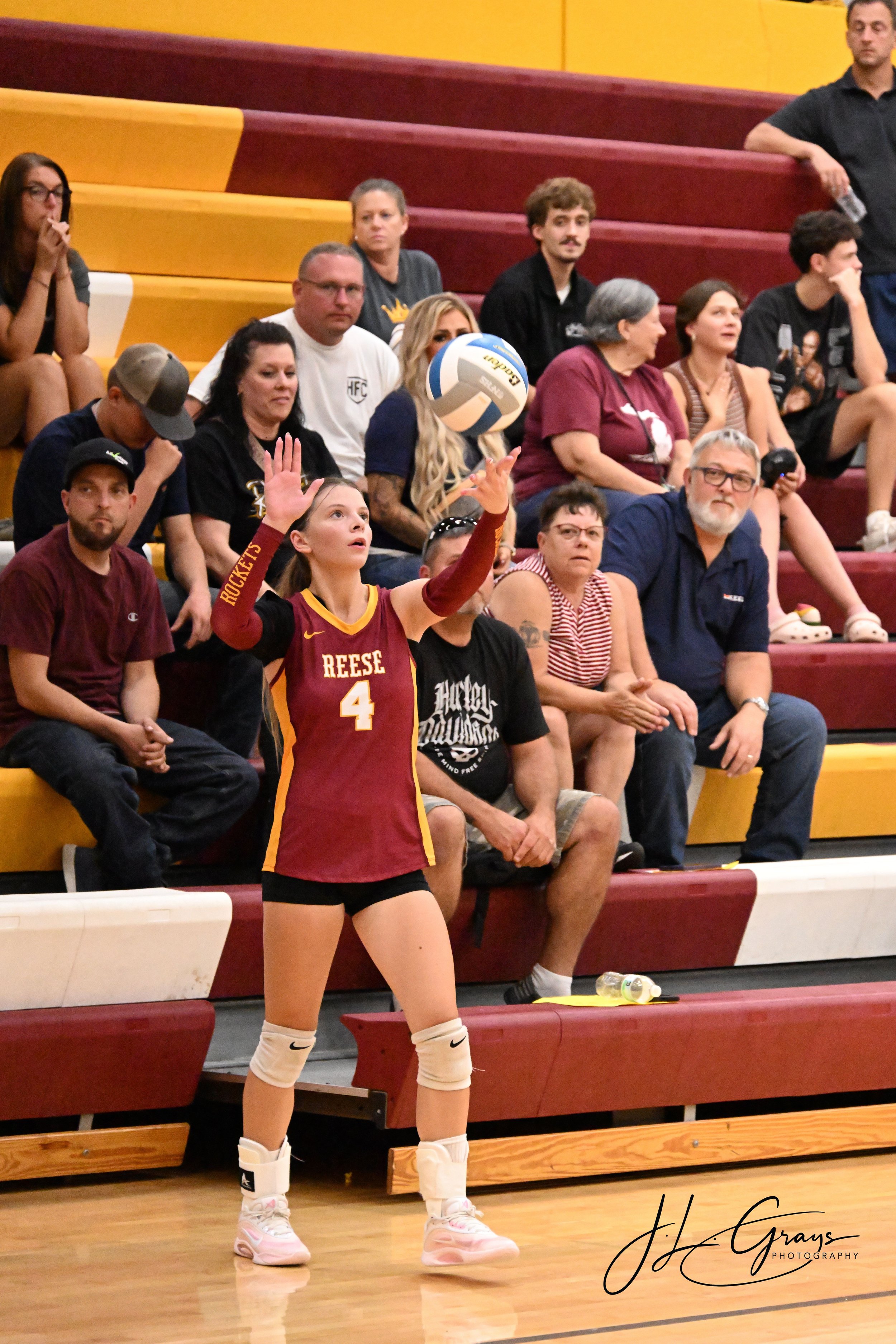 A female volleyball player in a red and yellow uniform preparing to serve in a gymnasium, with spectators seated behind her on red and yellow bleachers.