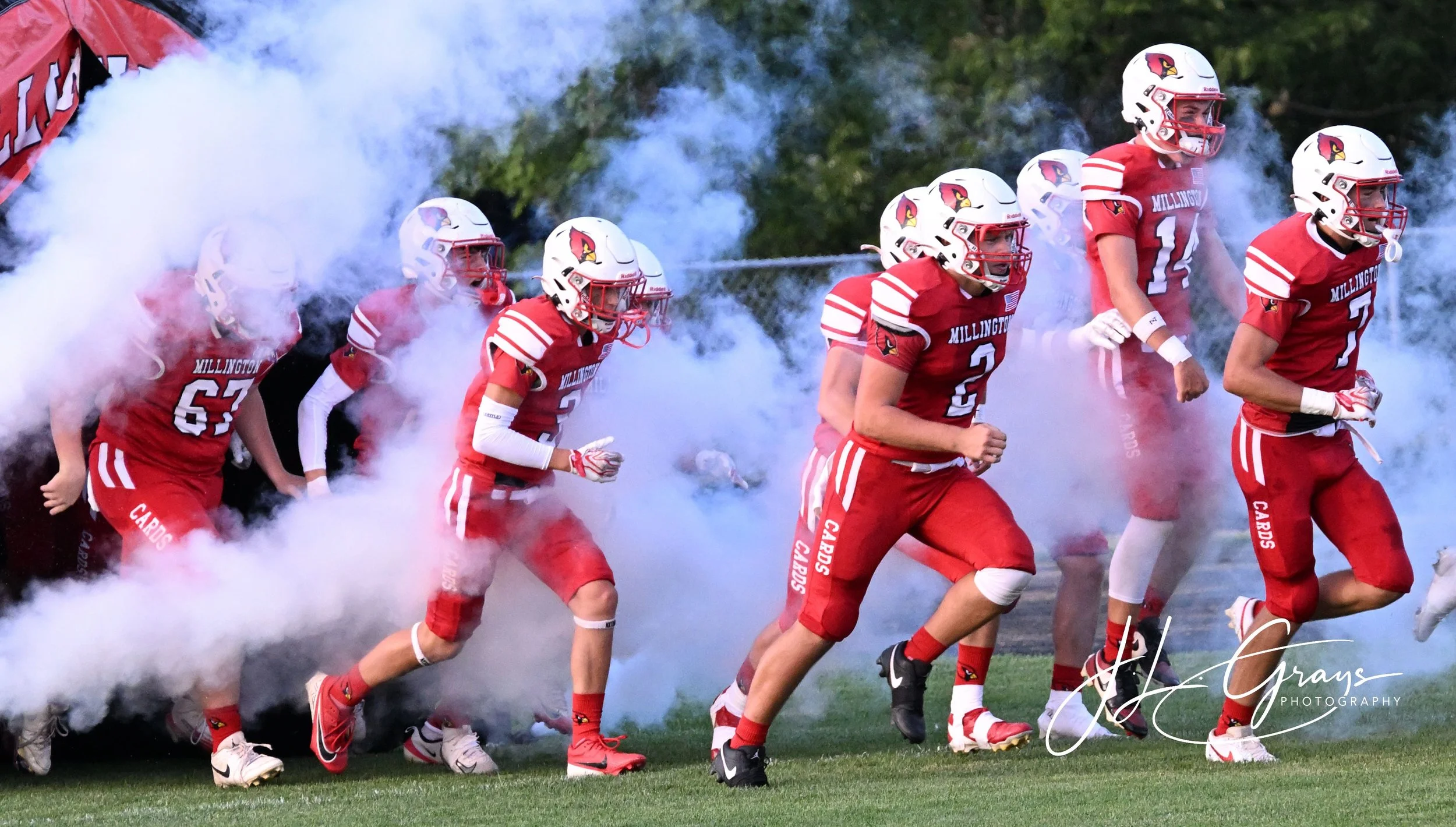 American football team in red uniforms and white helmets with a cardinal logo, running onto the field through smoke and led by a coach, during daytime.
