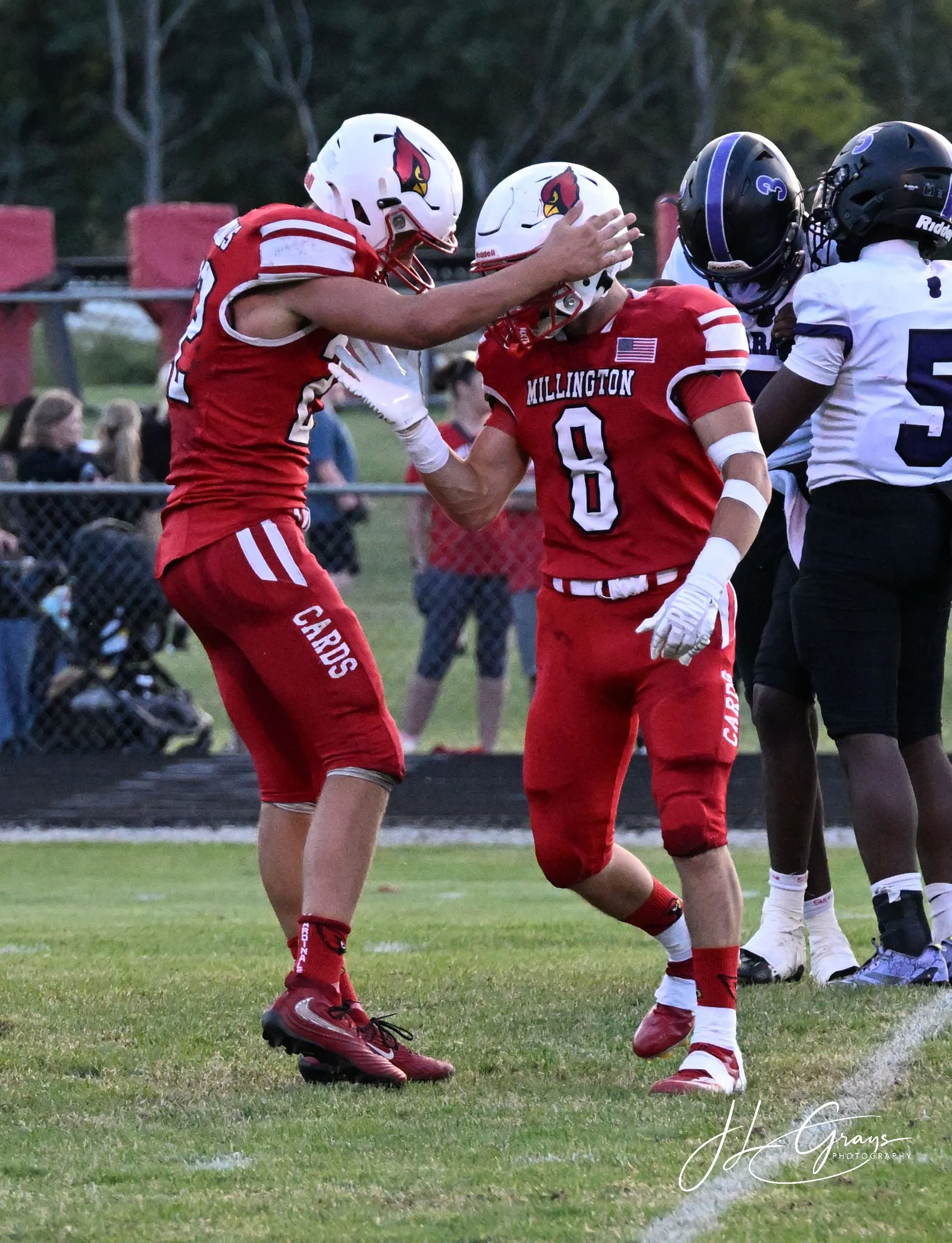 Two football players in red uniforms with white helmets sharing a moment on the field, with other players in the background, during a game.
