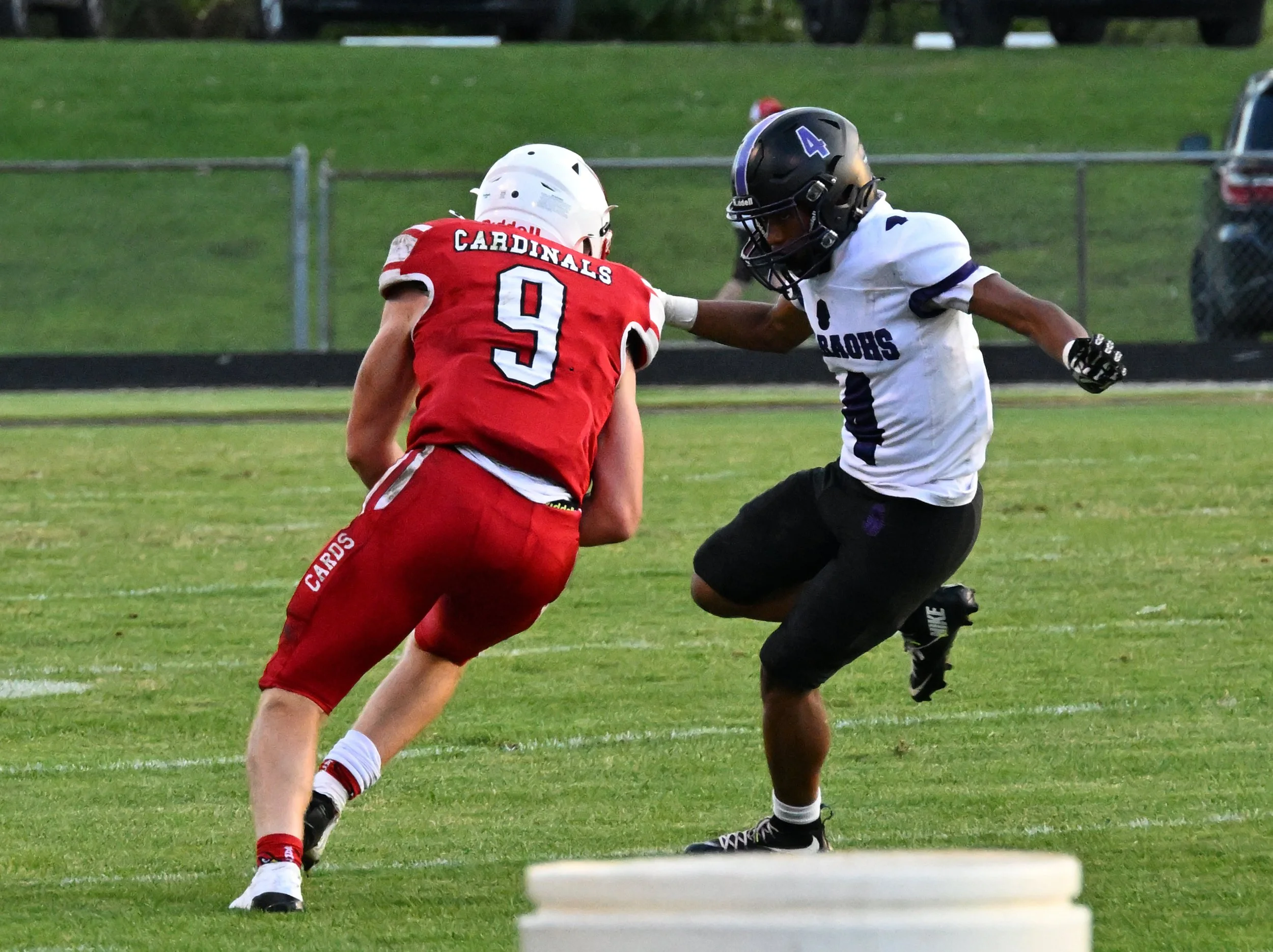 Two high school football players competing for the ball on the field. One player in a red uniform with number 9 and a white helmet facing away from the camera, the other in a white uniform with purple accents and number 4, wearing a black helmet, reaching out toward the player in red.