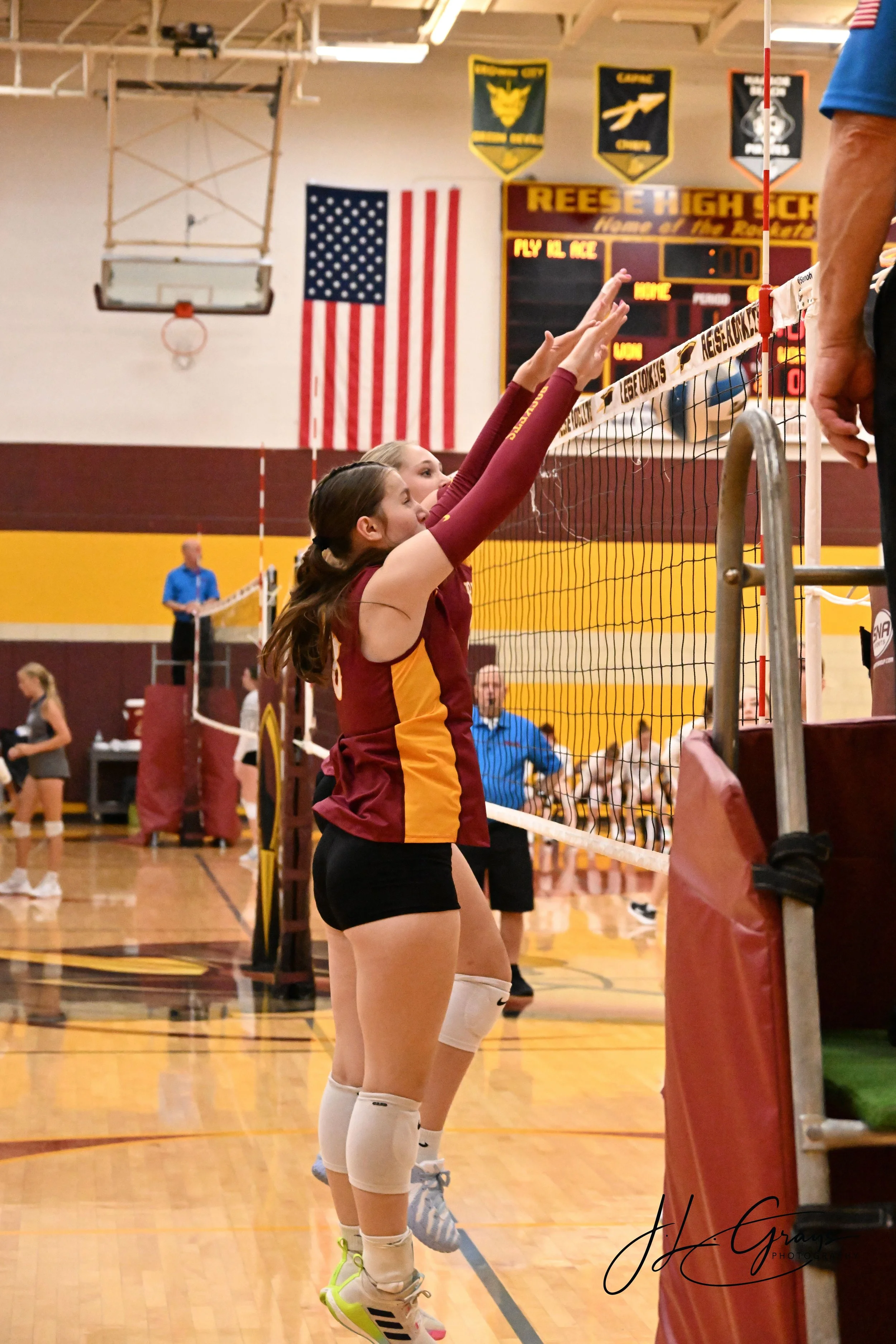 Two female volleyball players in maroon and gold uniforms reach above their heads to block a ball at the net during a volleyball game in a gymnasium. The gym has a wooden floor, a basketball hoop, and an American flag hanging on the wall. A scoreboar