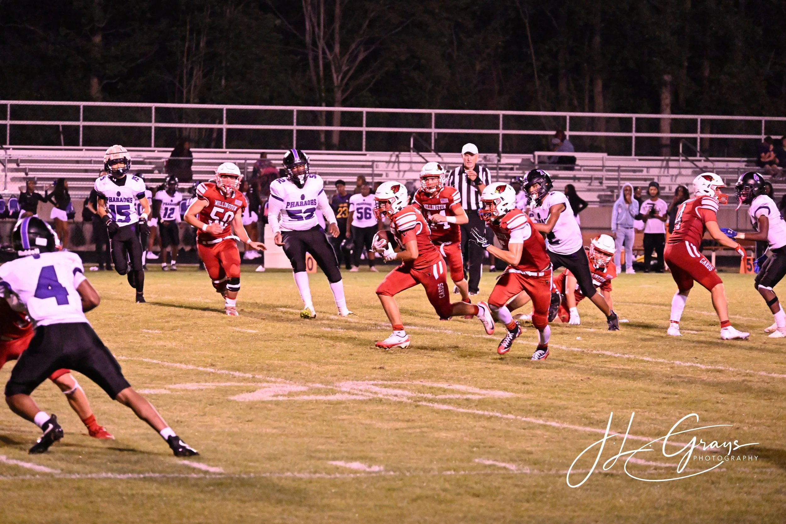 American football game at night with players in white and red uniforms on the field, a referee in black and white stripes, and spectators in the stands.