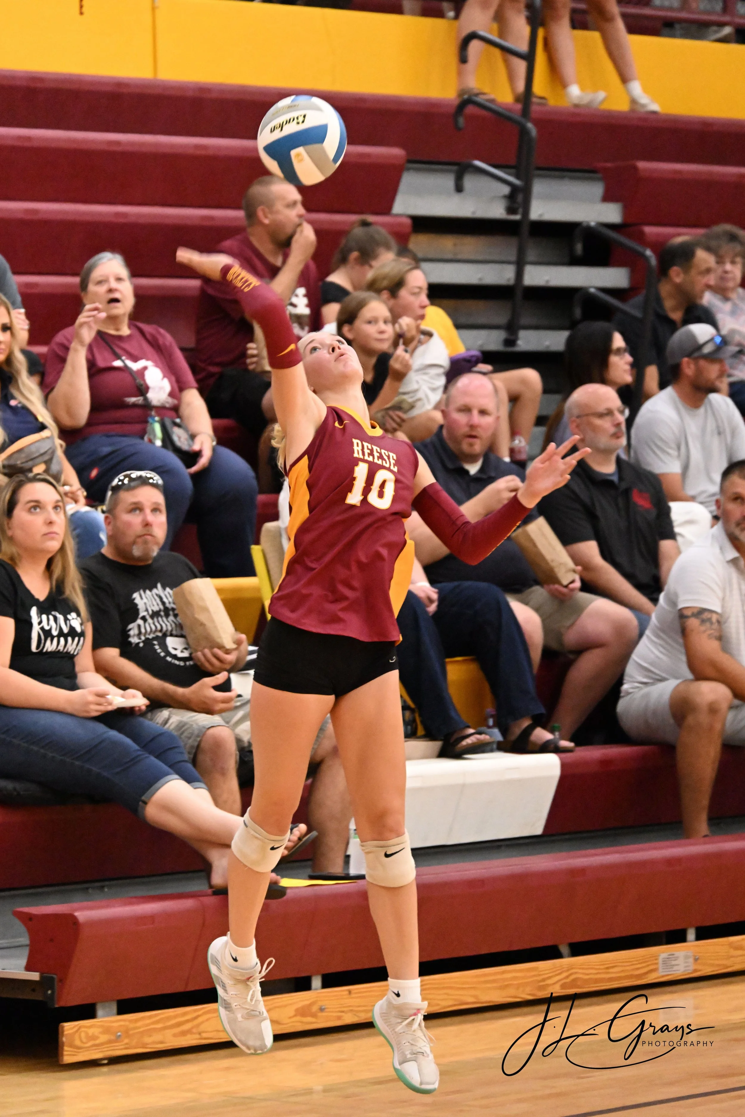 A female volleyball player in mid-air preparing to hit a volleyball during a game. She is wearing a maroon and yellow uniform with the number 10 and the name Reese on it, along with white Nike sneakers and knee pads. The background shows spectators s