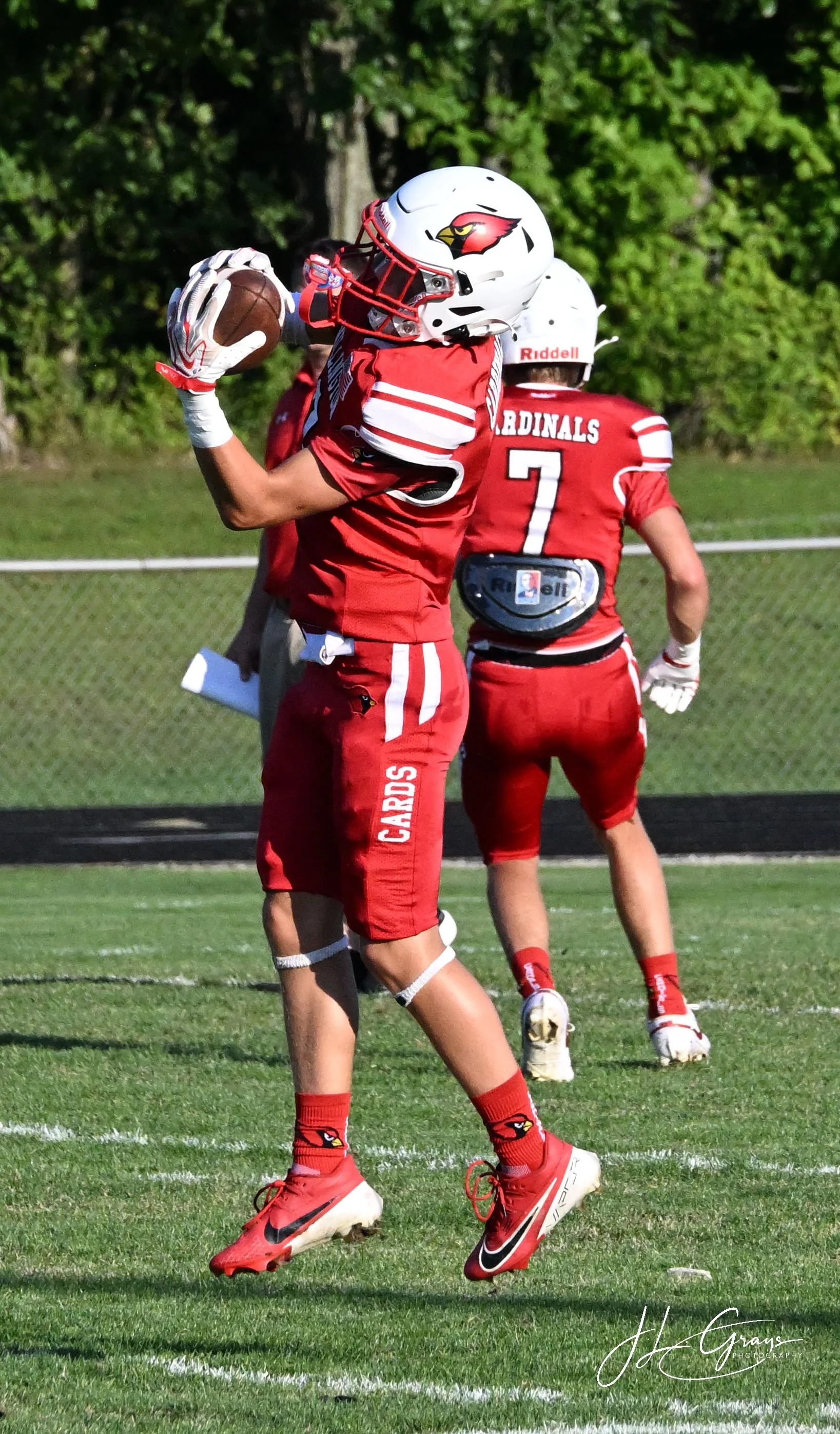 American football player in red uniform catching a football on a grass field with green trees in the background.