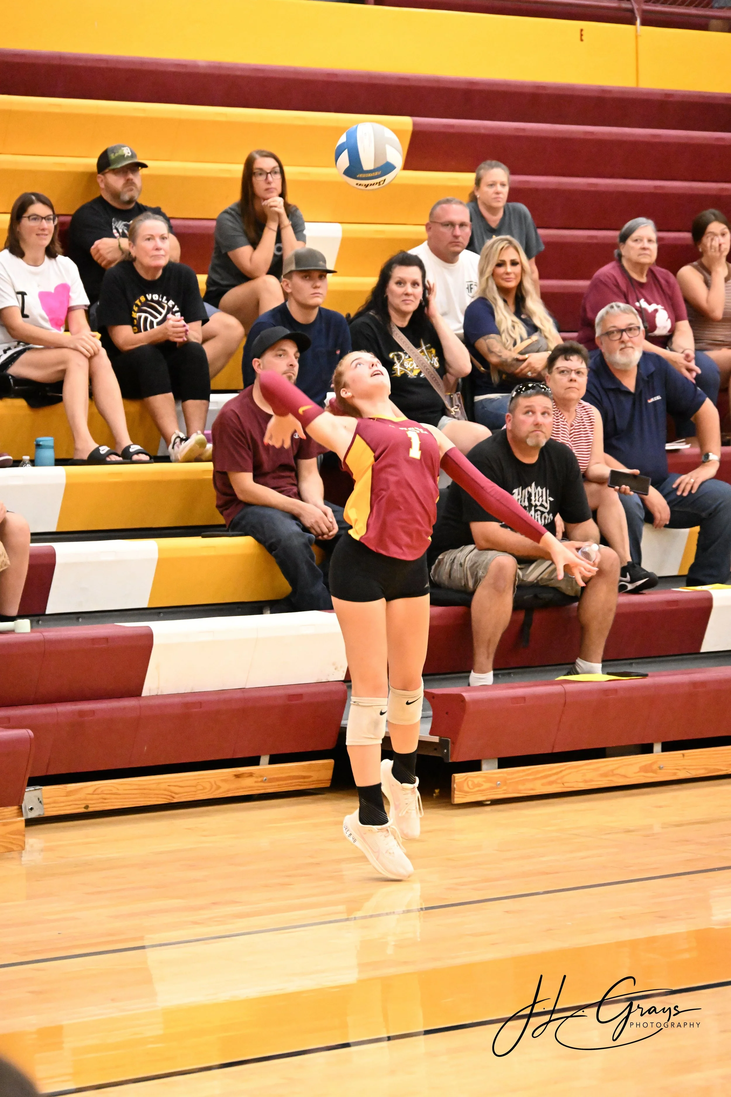 A volleyball player wearing a maroon and gold uniform with the number 1, standing on a gymnasium floor. Behind her, there are spectators sitting on yellow and maroon bleachers, watching a volleyball game.