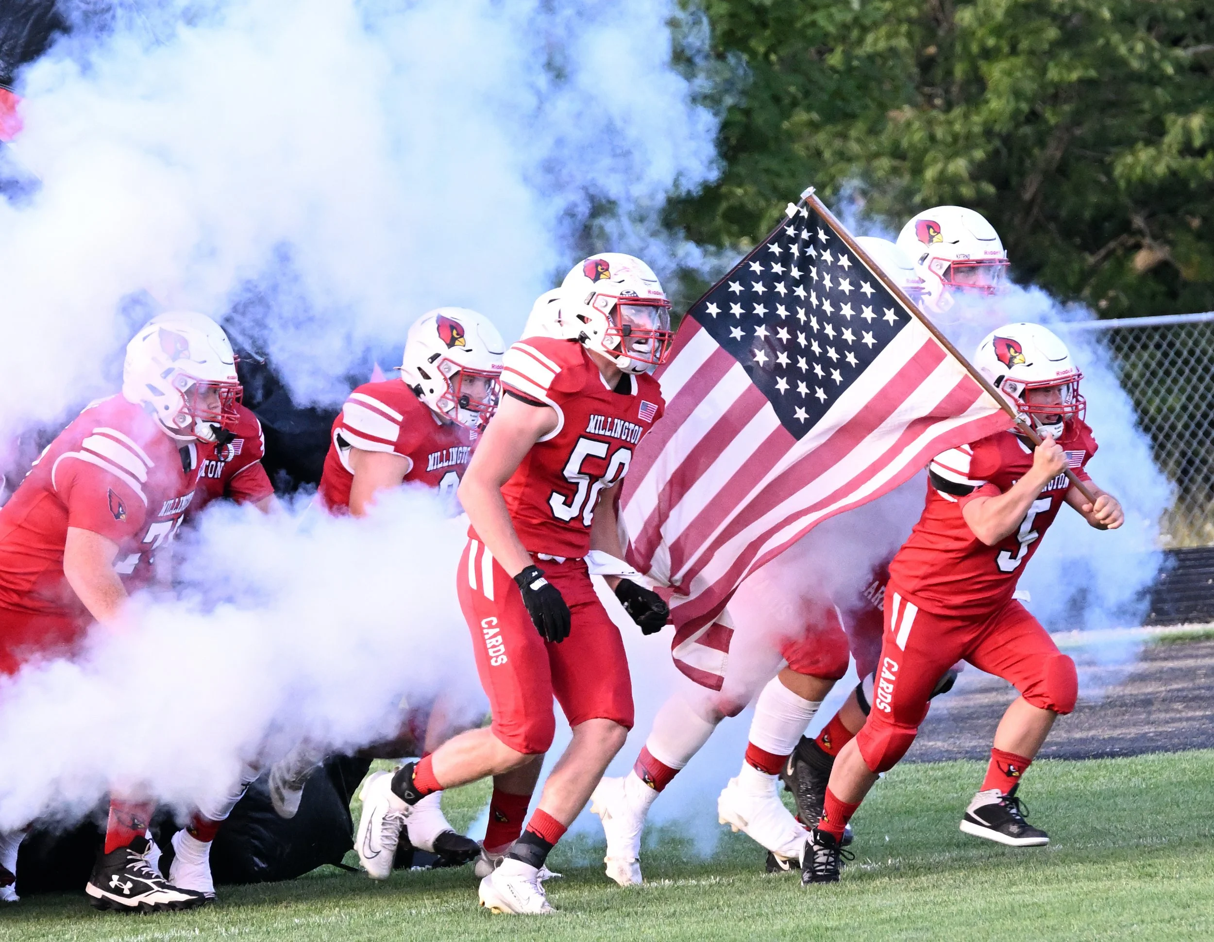 A group of football players in red uniforms and white helmets, some with the Cardinals logo, running onto the field through a cloud of smoke. One player is holding an American flag.