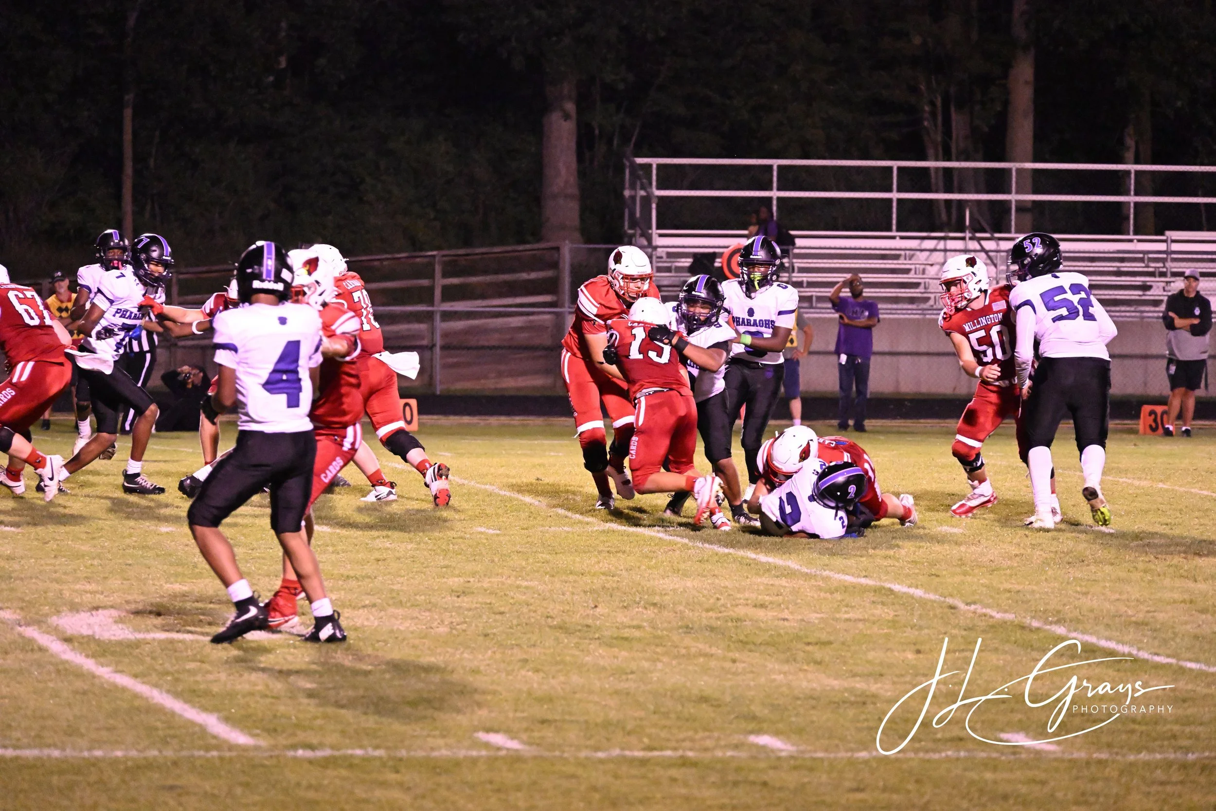 Young football players in red and white uniforms playing on a grass field at night, with some players on the ground and others standing, surrounded by empty bleachers and trees in the background.