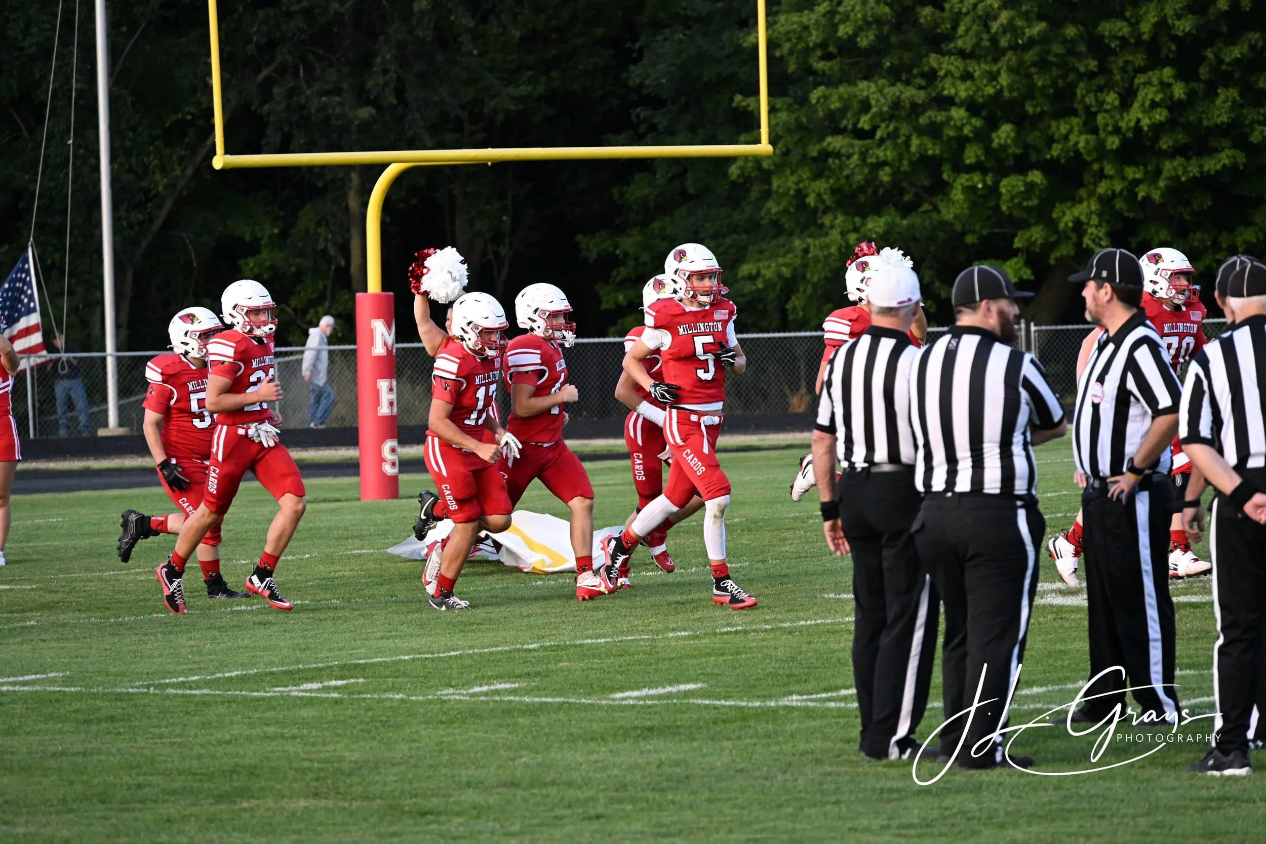 A group of American football players in red uniforms and helmets, some cheerleaders with pom-poms, and referees in black and white striped shirts on a football field with a goalpost, trees, and spectators in the background.