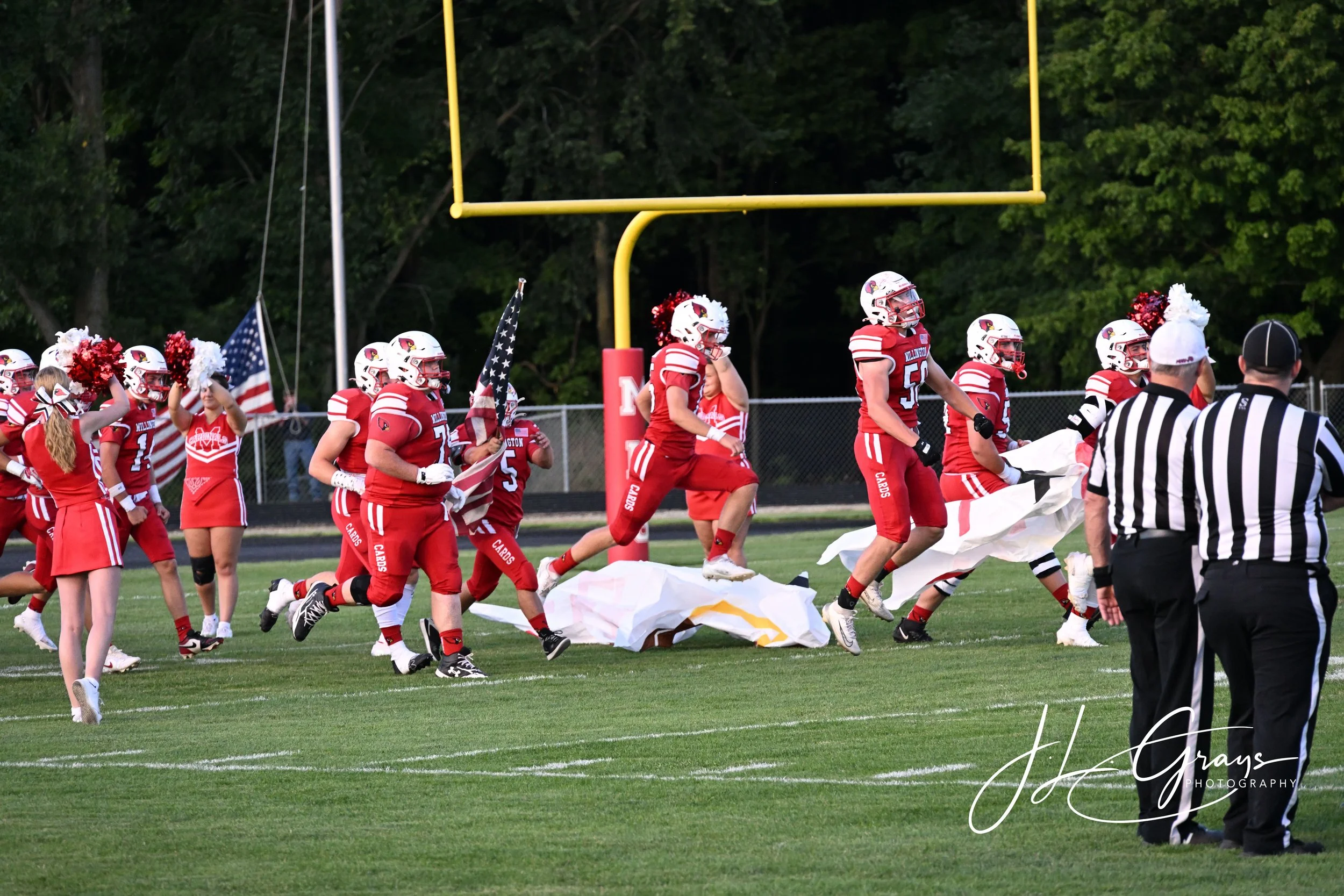 High school football players in red uniforms on the field with cheerleaders and an official, preparing for an event or game, near a goal post with a forest in the background.