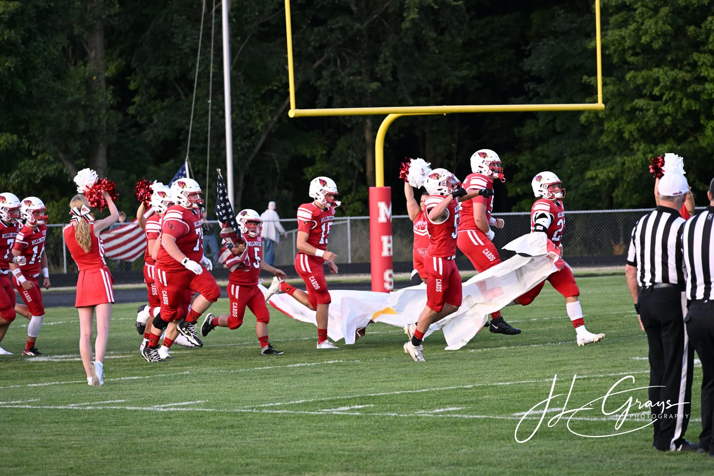 High school football players in red uniforms and white helmets on the field, participating in a team huddle or pre-game activity, with a football goalpost and trees in the background.