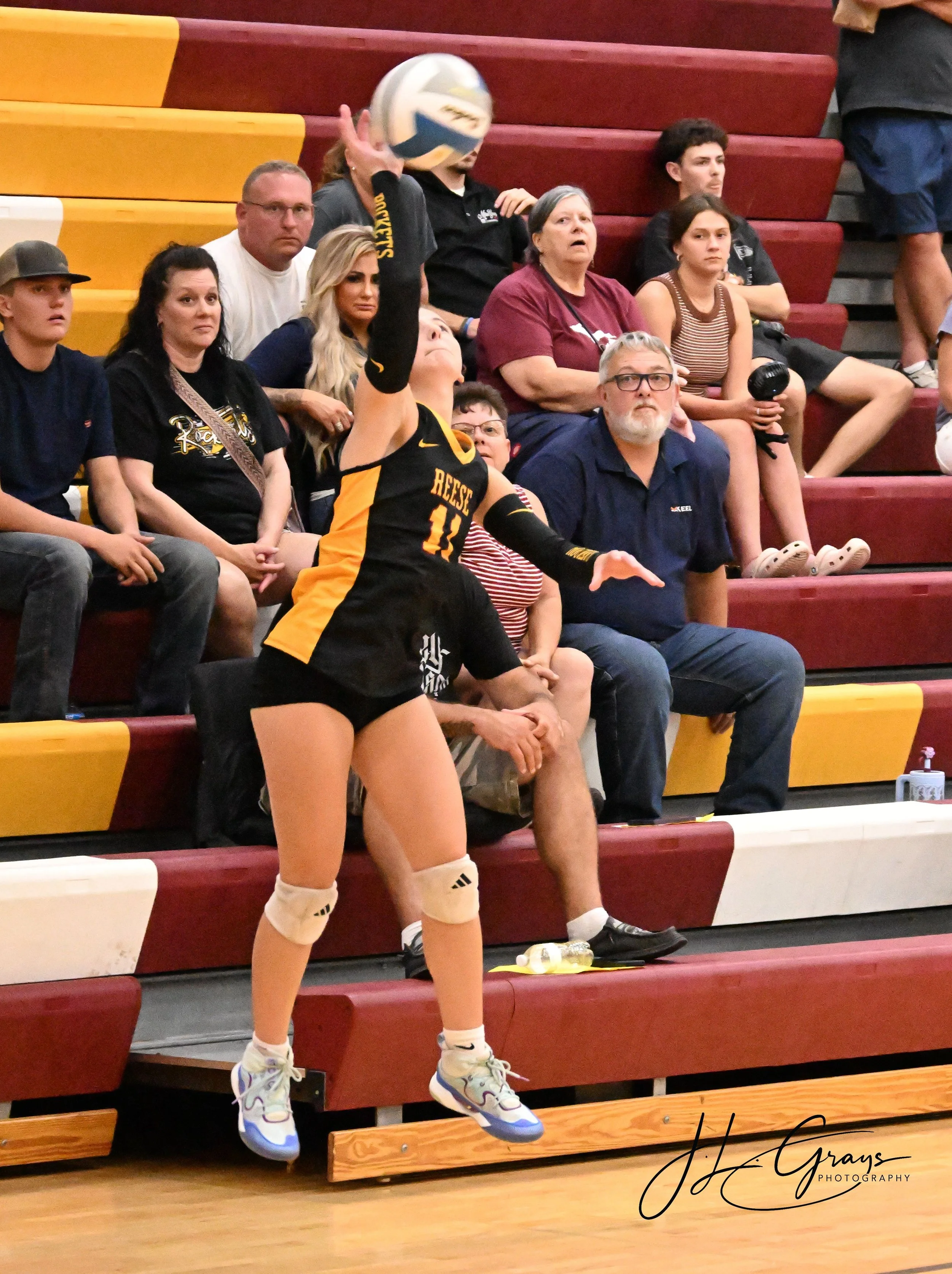 A female volleyball player with blonde hair wearing volleyball gear, including knee pads and sneakers, is in the foreground, jumping to serve the ball during a match. Spectators are seated on red and yellow bleachers in the background, watching the g