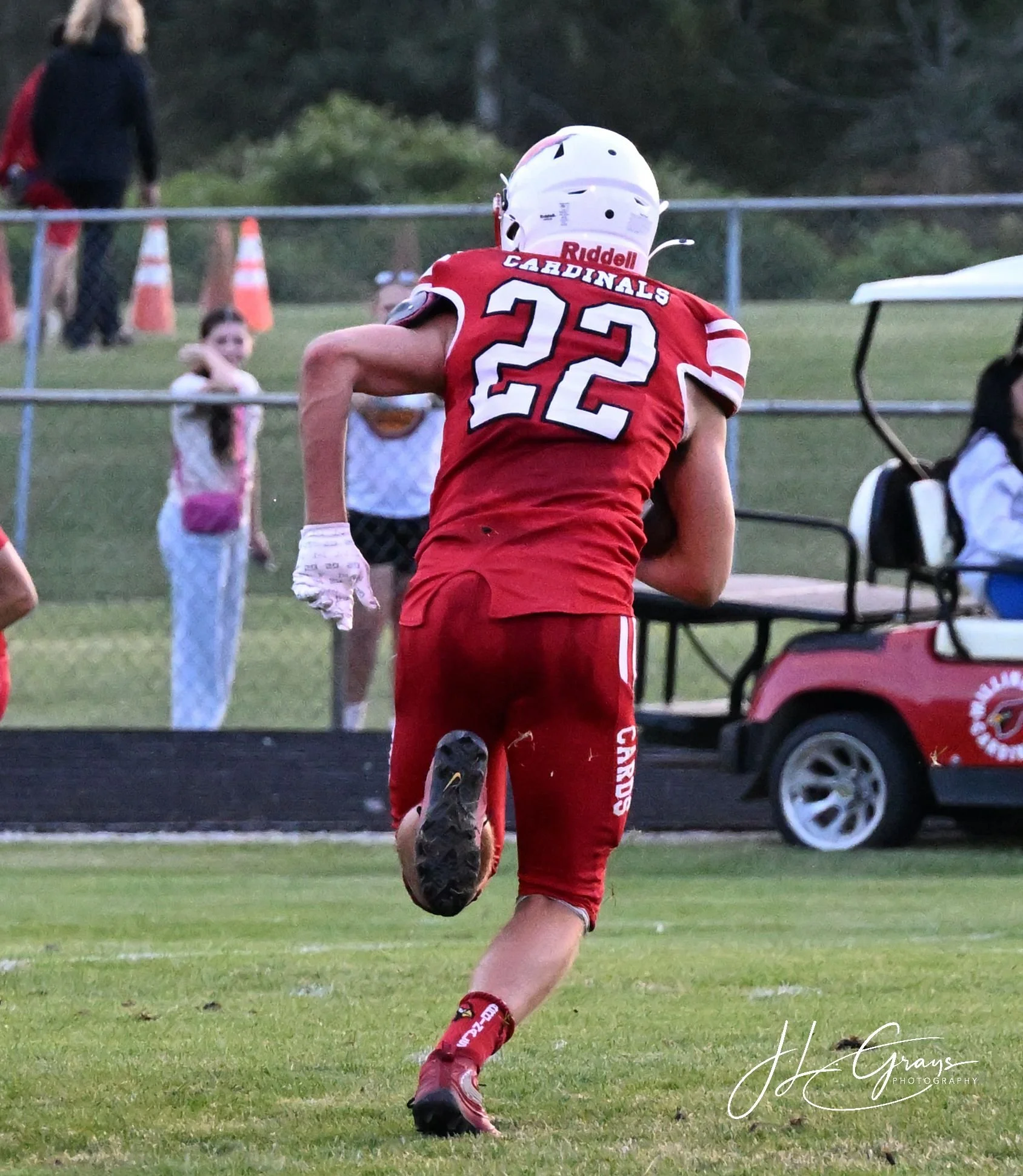A football player wearing an Atlanta Cardinals uniform, number 22, running on the field during a game. The player is seen from behind, with a white helmet and red uniform. In the background, there are children and adults, some standing near the sidel
