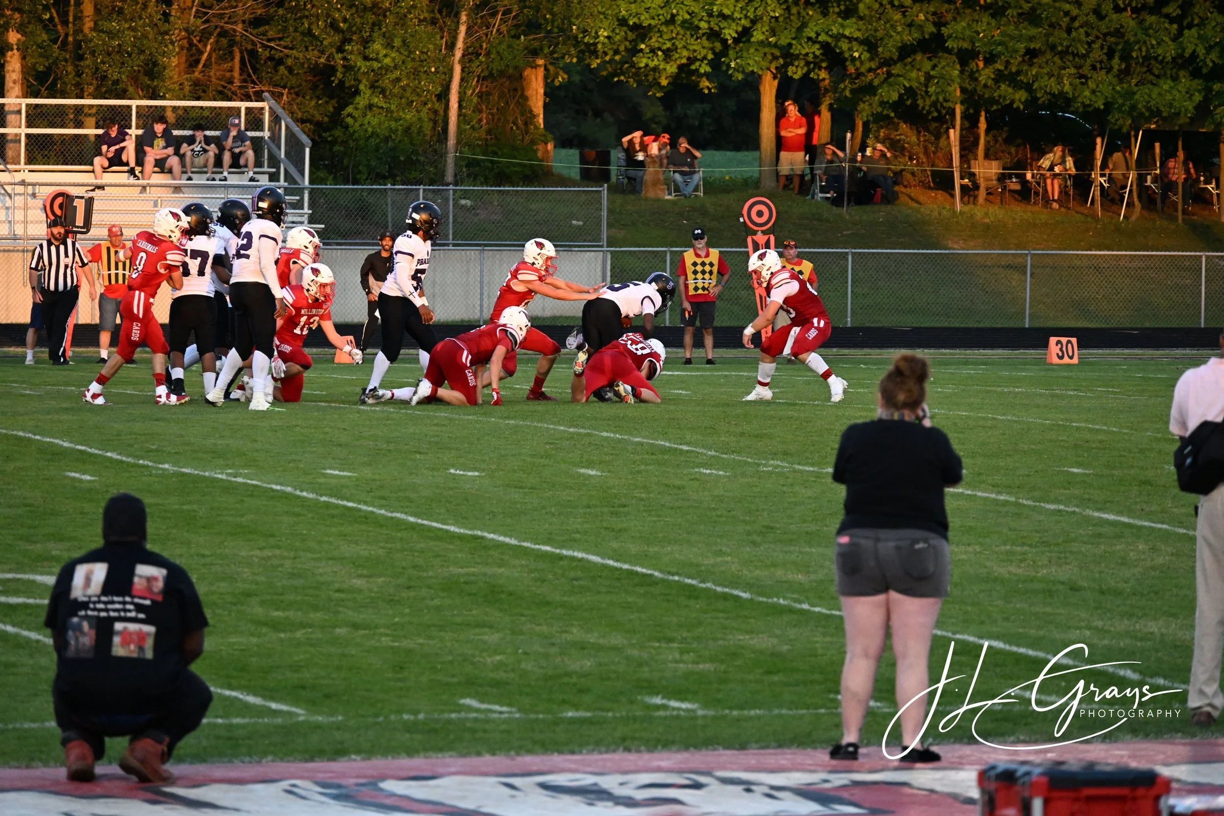 A football game is in action on the field with players in red and white uniforms and players in black and white uniforms. Players are engaged in a play, with some on the ground and others preparing for the next move. Spectators are watching from the 
