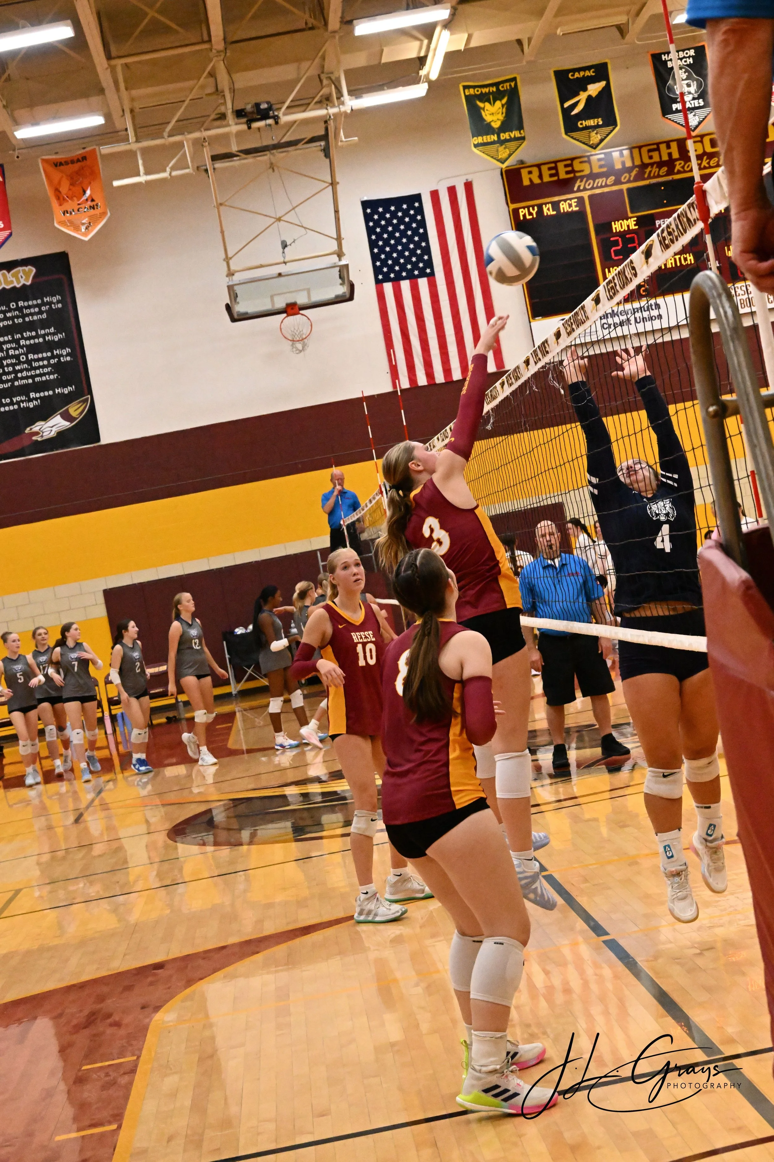 A volleyball game in progress at an indoor gymnasium. Female players are positioned near the net, with one jumping to spike the ball. The gym features banners on the wall, an American flag, and a scoreboard showing the score as 23-22. Spectators and 