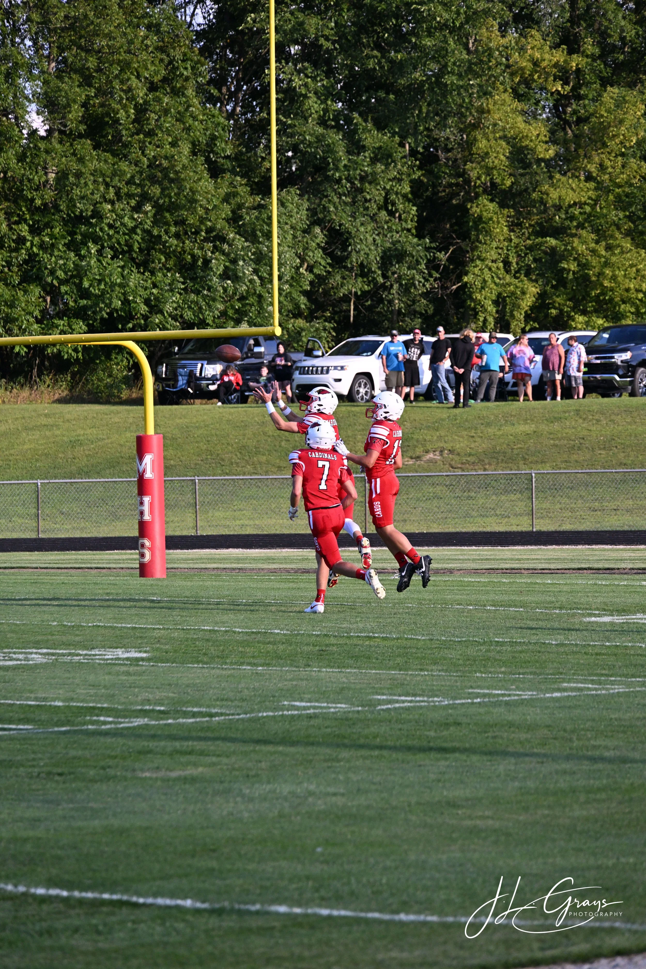 Football players in red uniforms playing on a field, with a goal post, and a crowd of spectators and parked cars in the background.