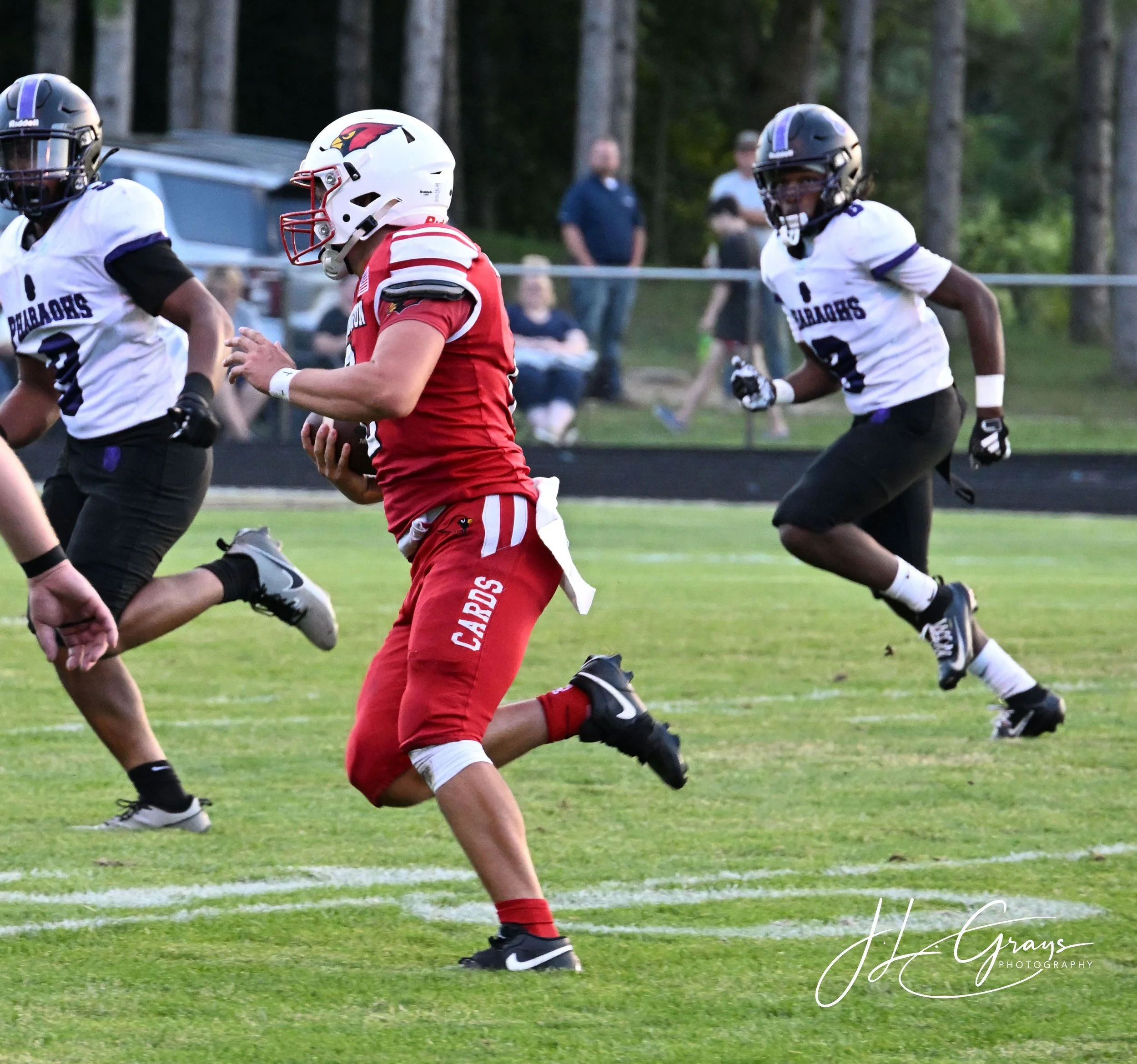 A football player in a red uniform with a helmet running with the ball, while two players in white jerseys and black helmets chase him on a grassy field in the woods.