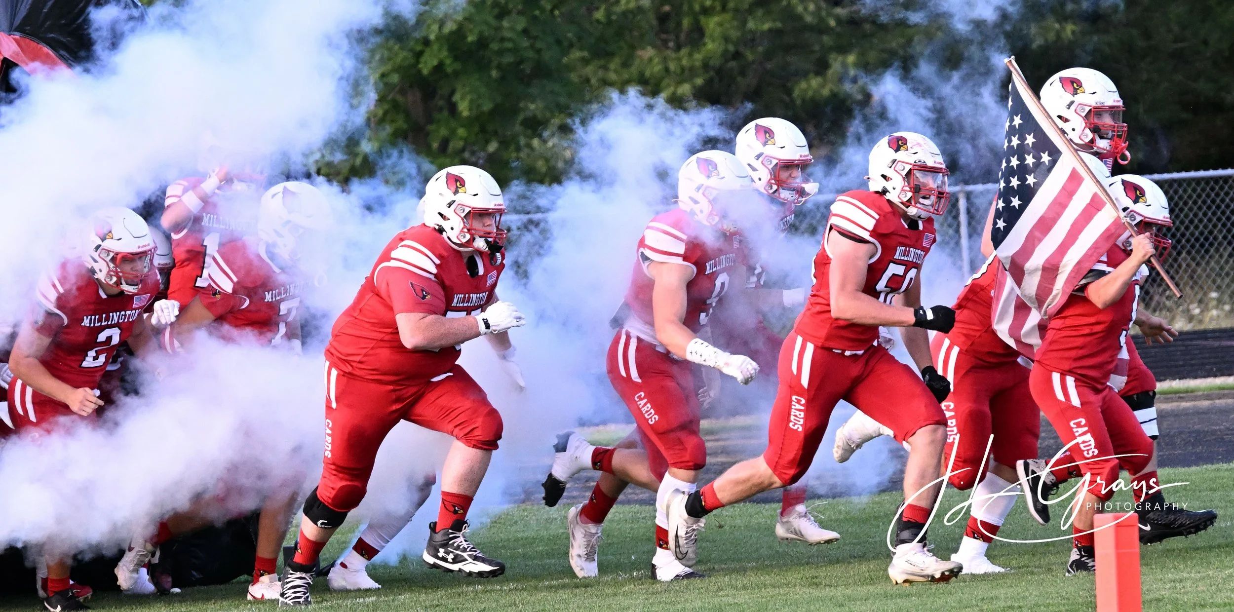 High school football players in red uniforms and helmets running onto the field through a cloud of smoke, with one player holding an American flag.