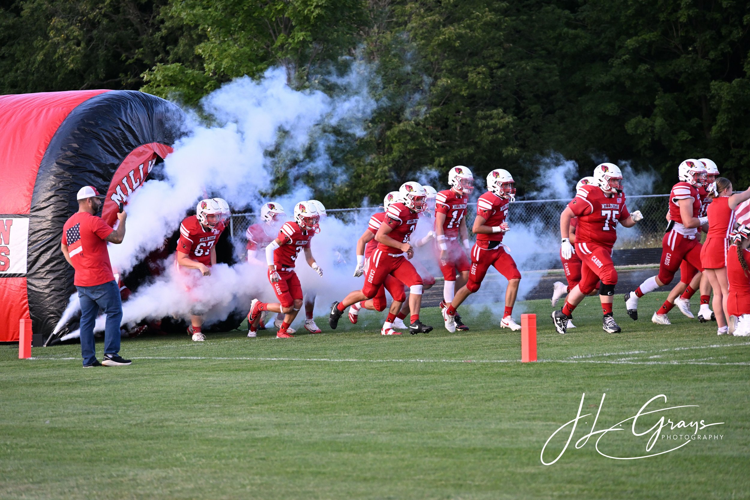 A football team in red and white uniforms runs onto the field through a smoke cloud from a large inflatable tunnel with the word 'Millington' on it. A person in a red shirt and white cap stands nearby, and green trees are visible in the background.