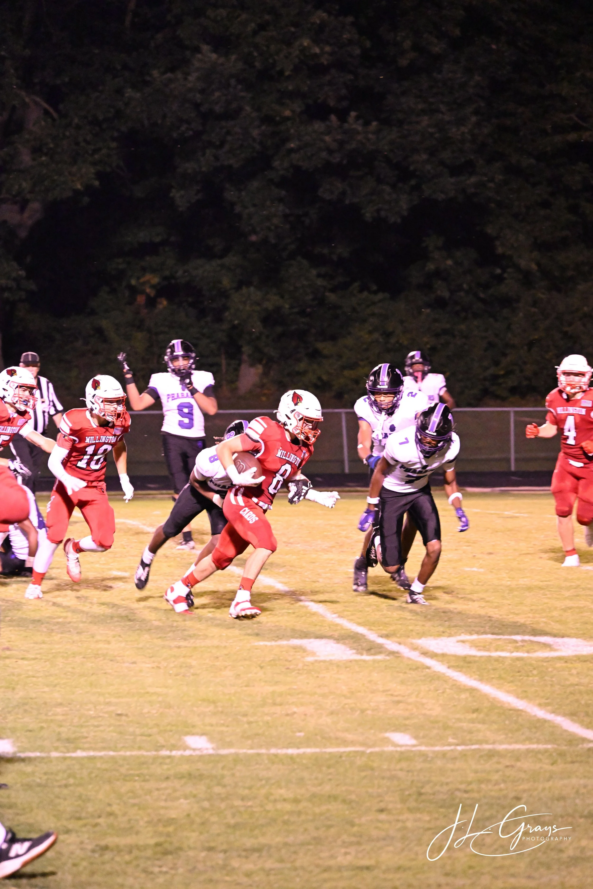 A football game is in progress on a night game field, with players in red and white uniforms and black and white uniforms. A player in a red uniform is running with the football, while other players are engaged in blocking or attempting to tackle. Th