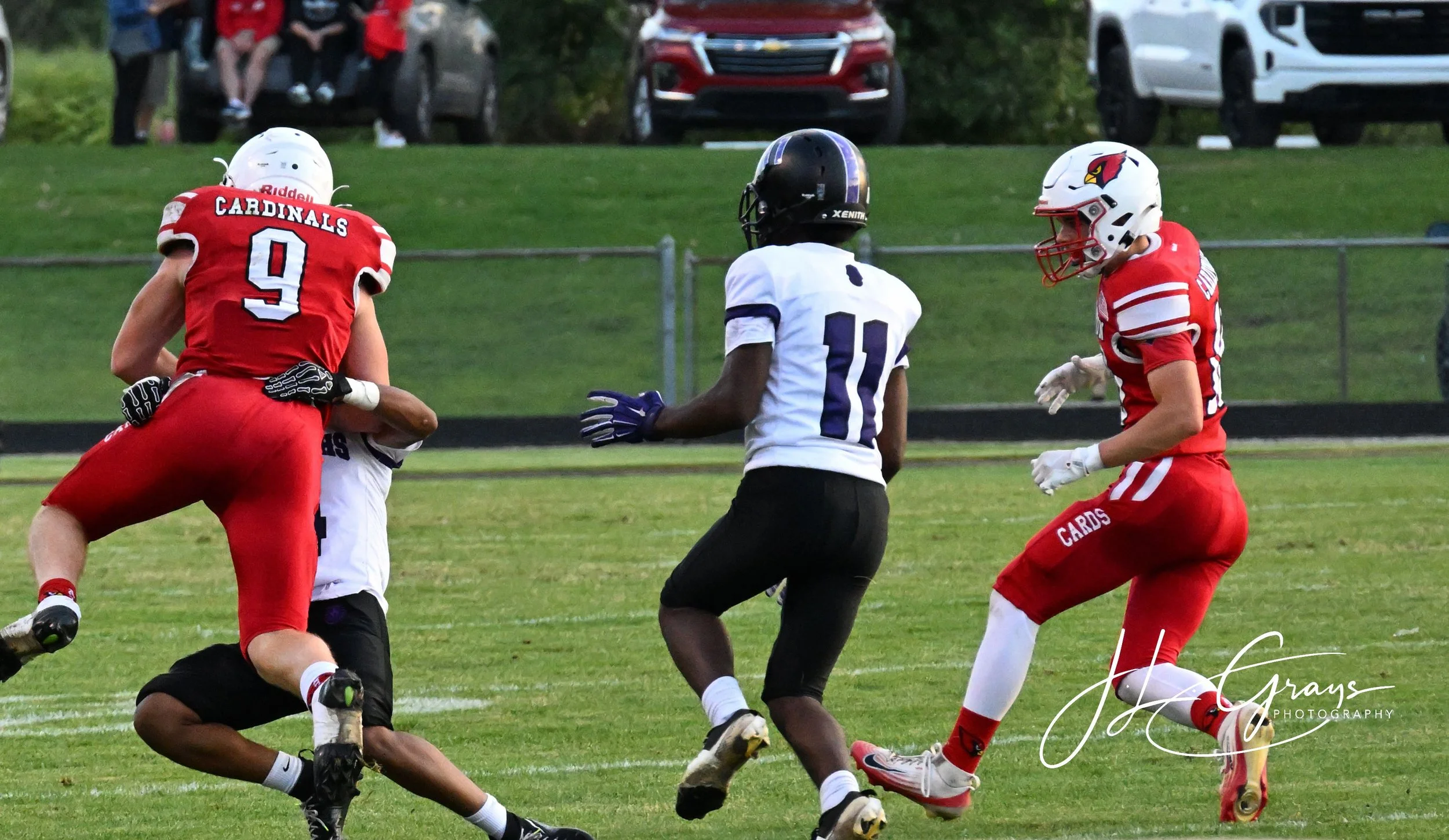 Youth football game with players in red and white uniforms and a player in black, on a grassy field with vehicles in the background.