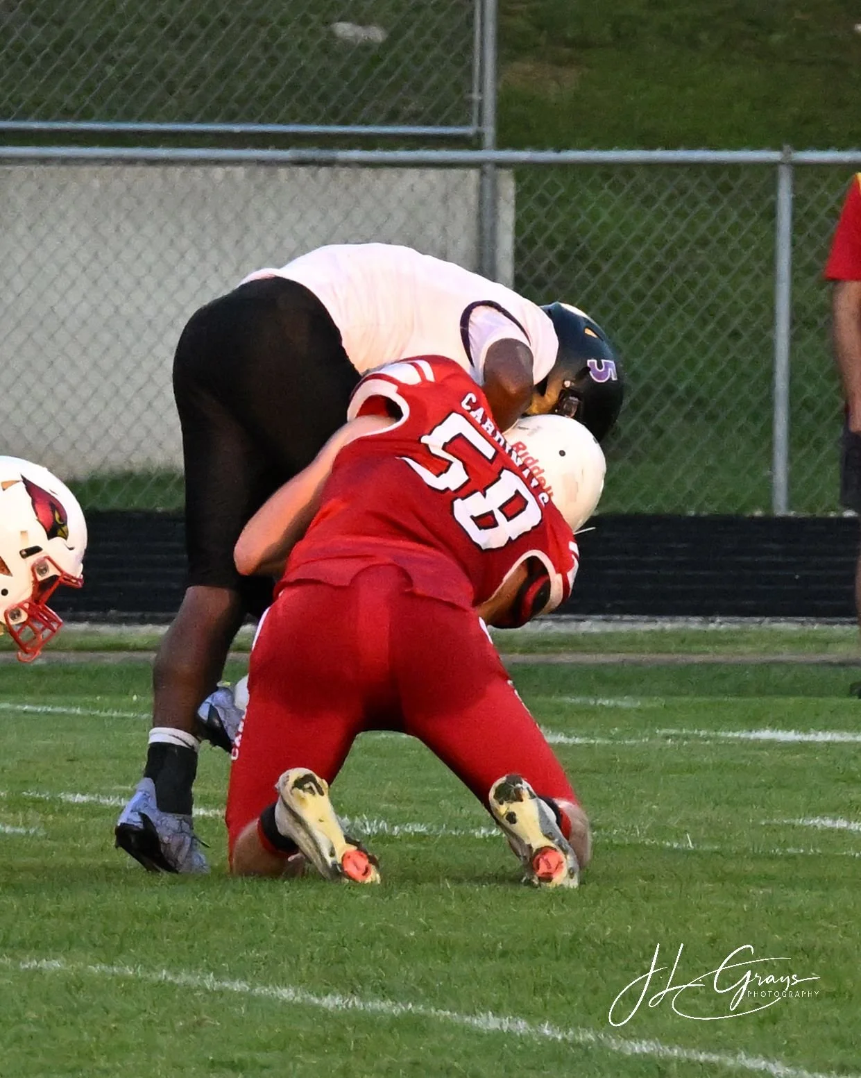 Two football players in a tackle during a game, one in a white jersey and the other in a red jersey, on a grass field with a chain-link fence in the background.