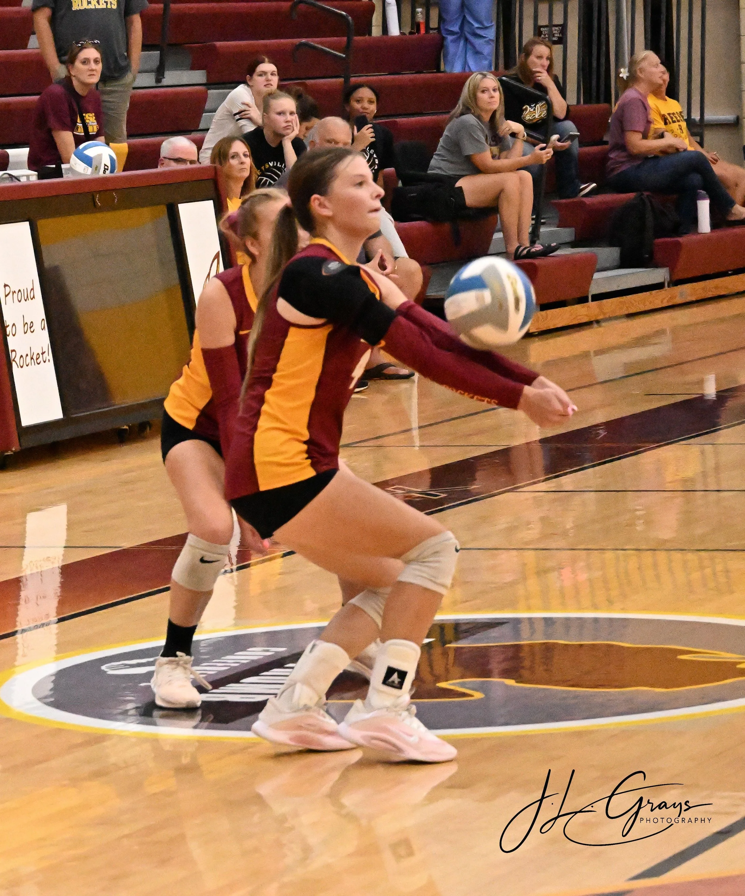 A female volleyball player in a maroon and yellow uniform serves the ball during a match inside a gymnasium, with spectators watching from the bleachers in the background.
