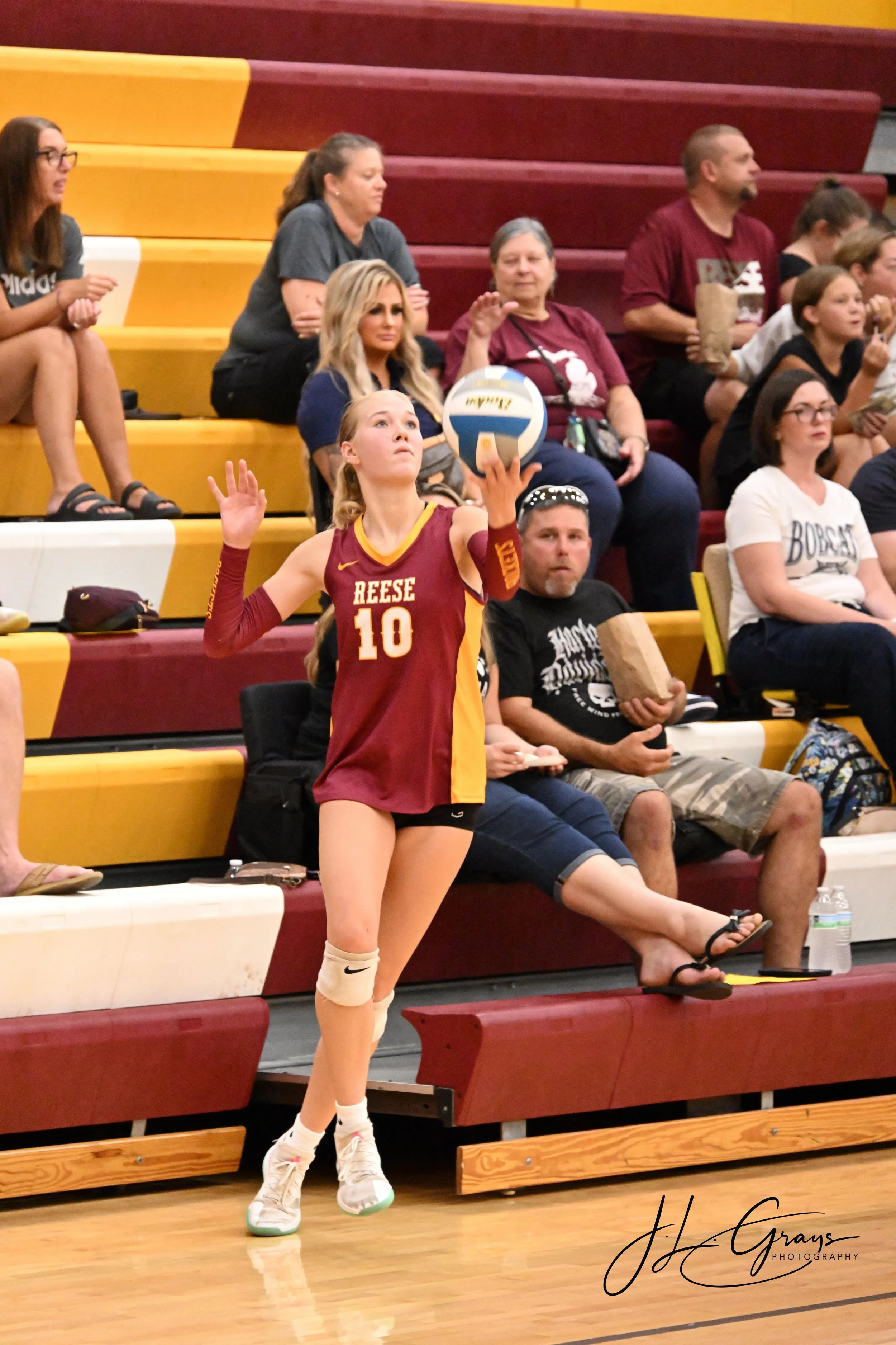 A young female volleyball player in a maroon and yellow uniform with the number 10, holding a volleyball in her right hand, standing on a gym floor with beige knee pads, white socks, and sneakers, with audience members seated in the background on red