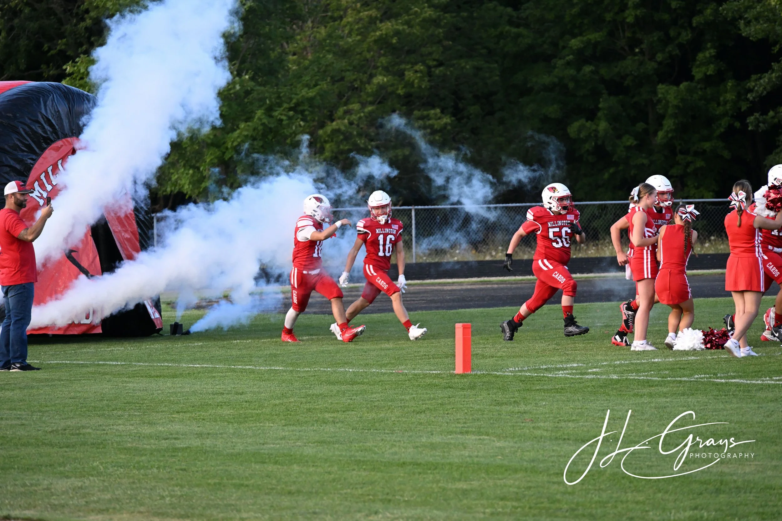 A group of football players in red uniforms and white helmets running onto a field through a smoke tunnel at a football game. There are cheerleaders in red and white outfits and a man taking a photo on the side. The scene is outdoors on a grassy fiel