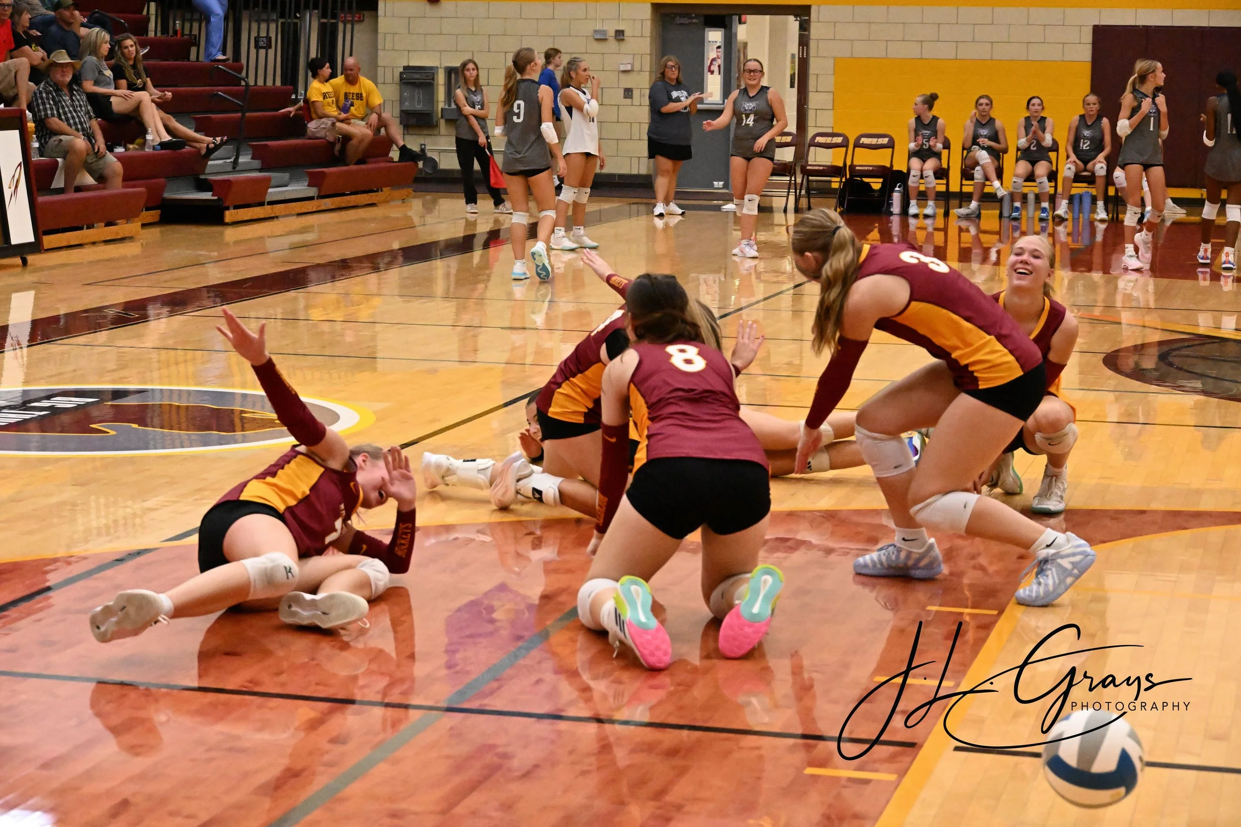 A volleyball game scene showing players in maroon and yellow uniforms celebrating on the gym floor, some lying and some kneeling, with volleyballs nearby. In the background, other players in gray uniforms stand, and fans and spectators sit on maroon 