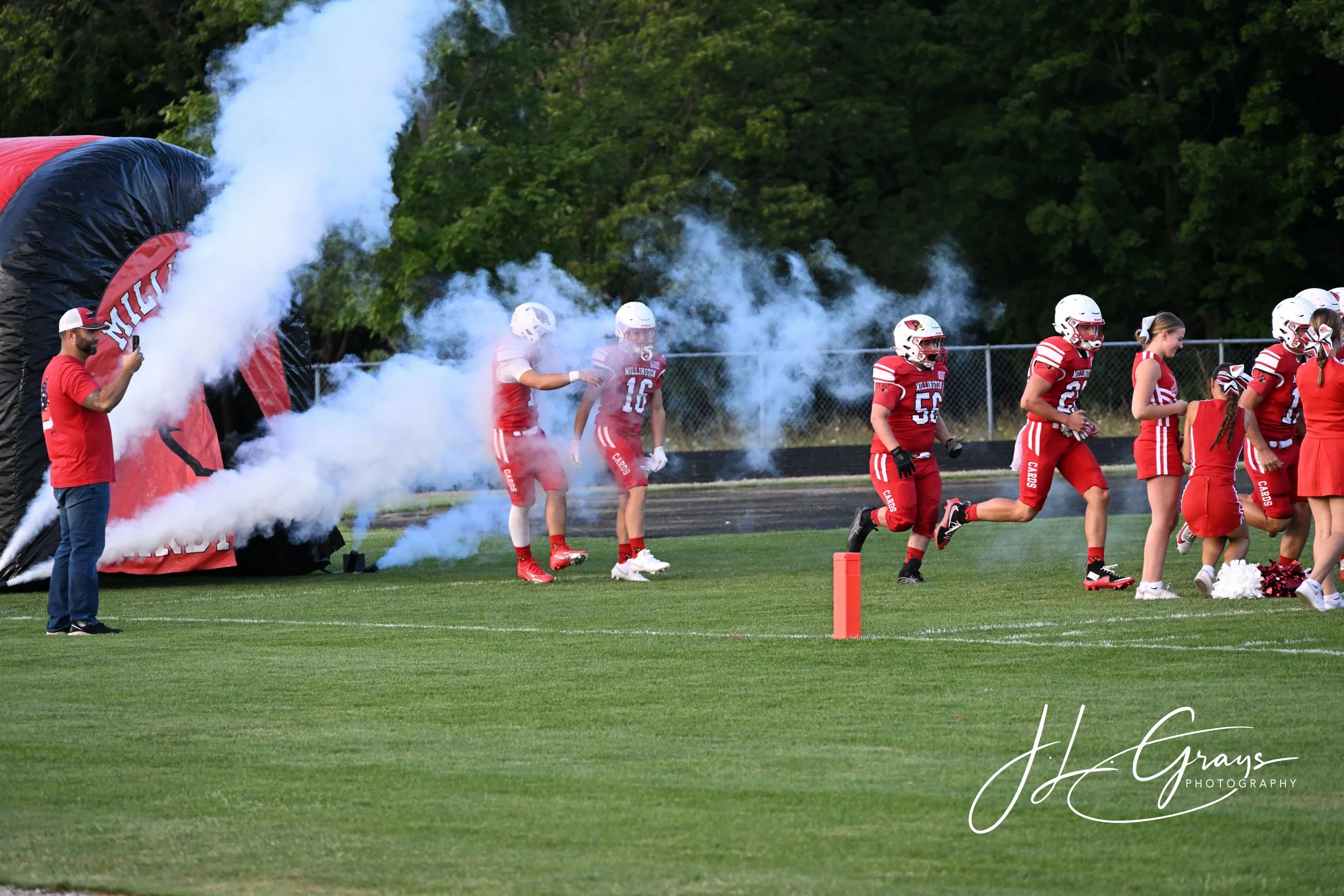 Football players in red uniforms running onto the field through a fog of smoke from a tunnel at a high school game, with a man taking a photo on his phone.