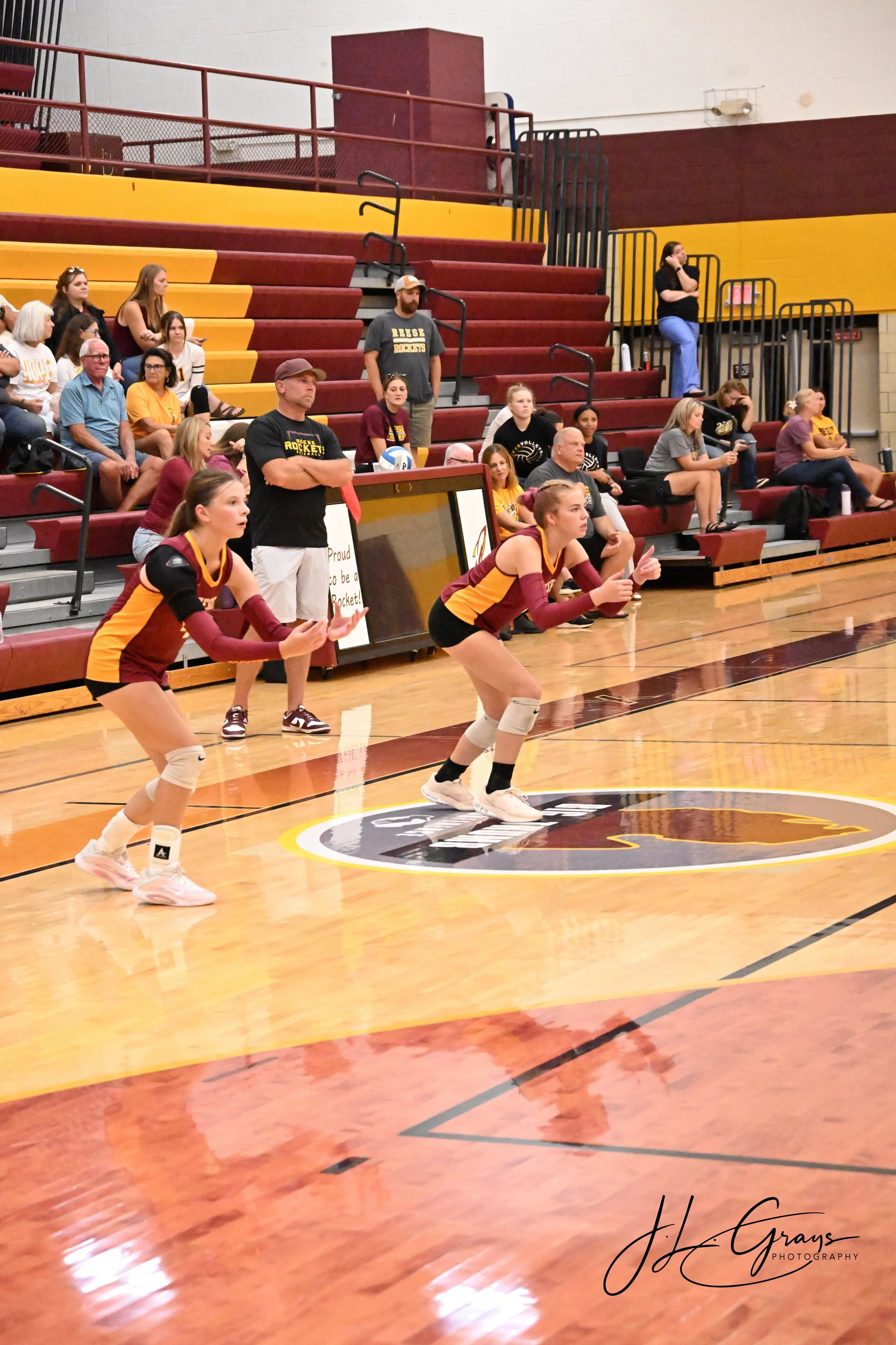 An indoor volleyball court with players in maroon and yellow jerseys, a coach, and spectators seated on tiered bleachers in the background.