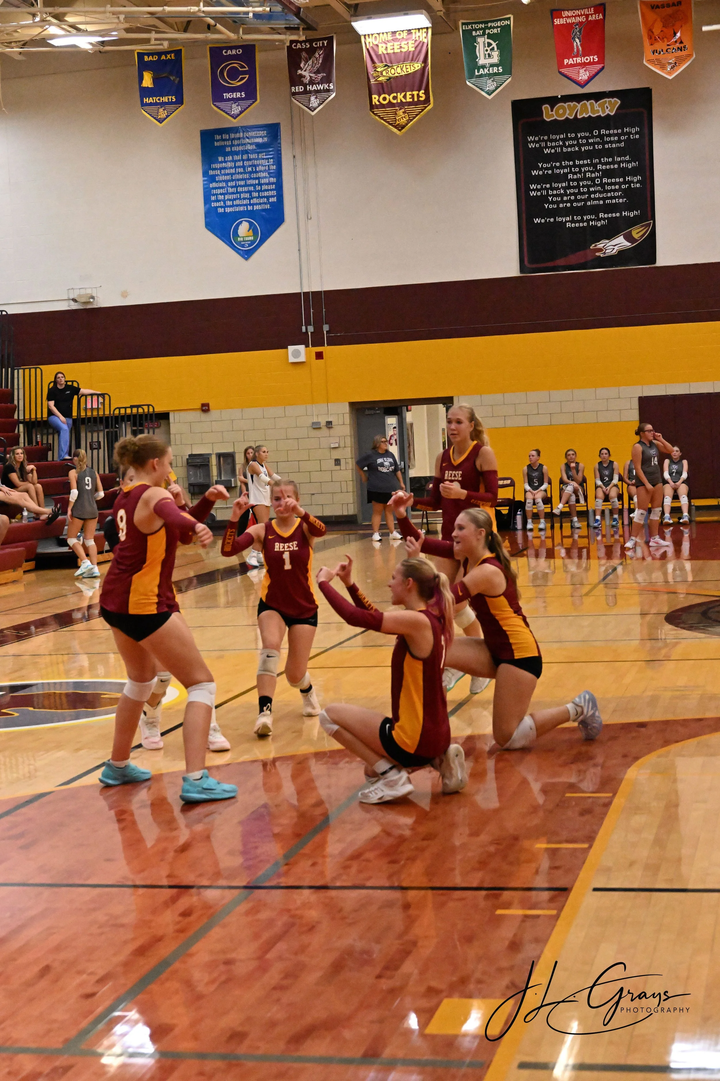 A girls' volleyball team in maroon and yellow uniforms celebrating a victory inside a gymnasium, with some players standing and others kneeling or sitting on the floor.