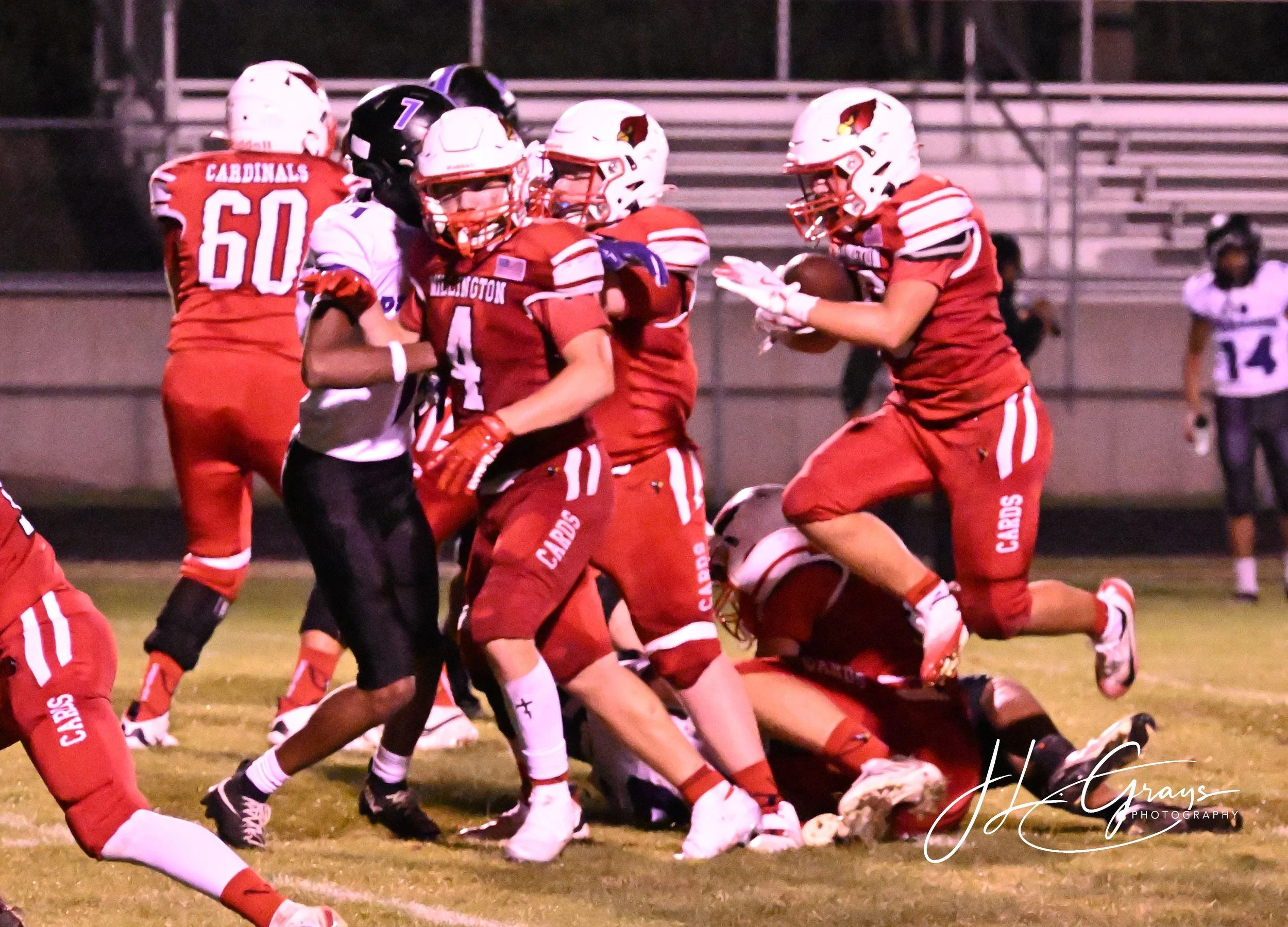 A group of football players in red and white uniforms and helmets are on the field during a game. One player is holding the football, while others are engaged in blocking or tackling. The scene captures action and teamwork.
