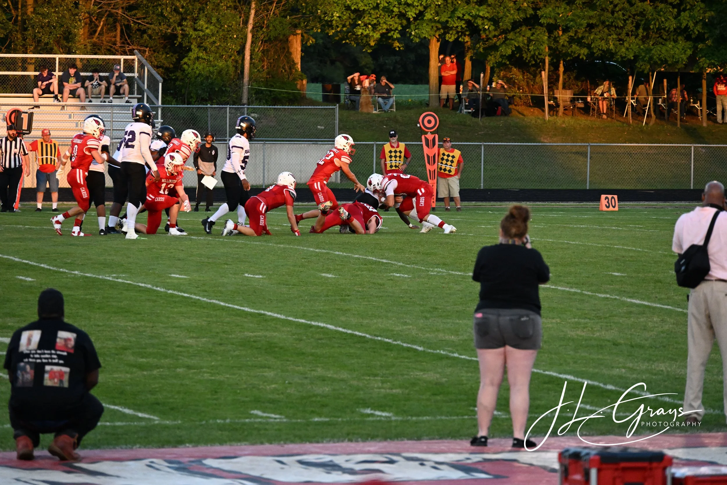 American football game in progress with players on the field and spectators in the stands and on the hill in the background during evening time.
